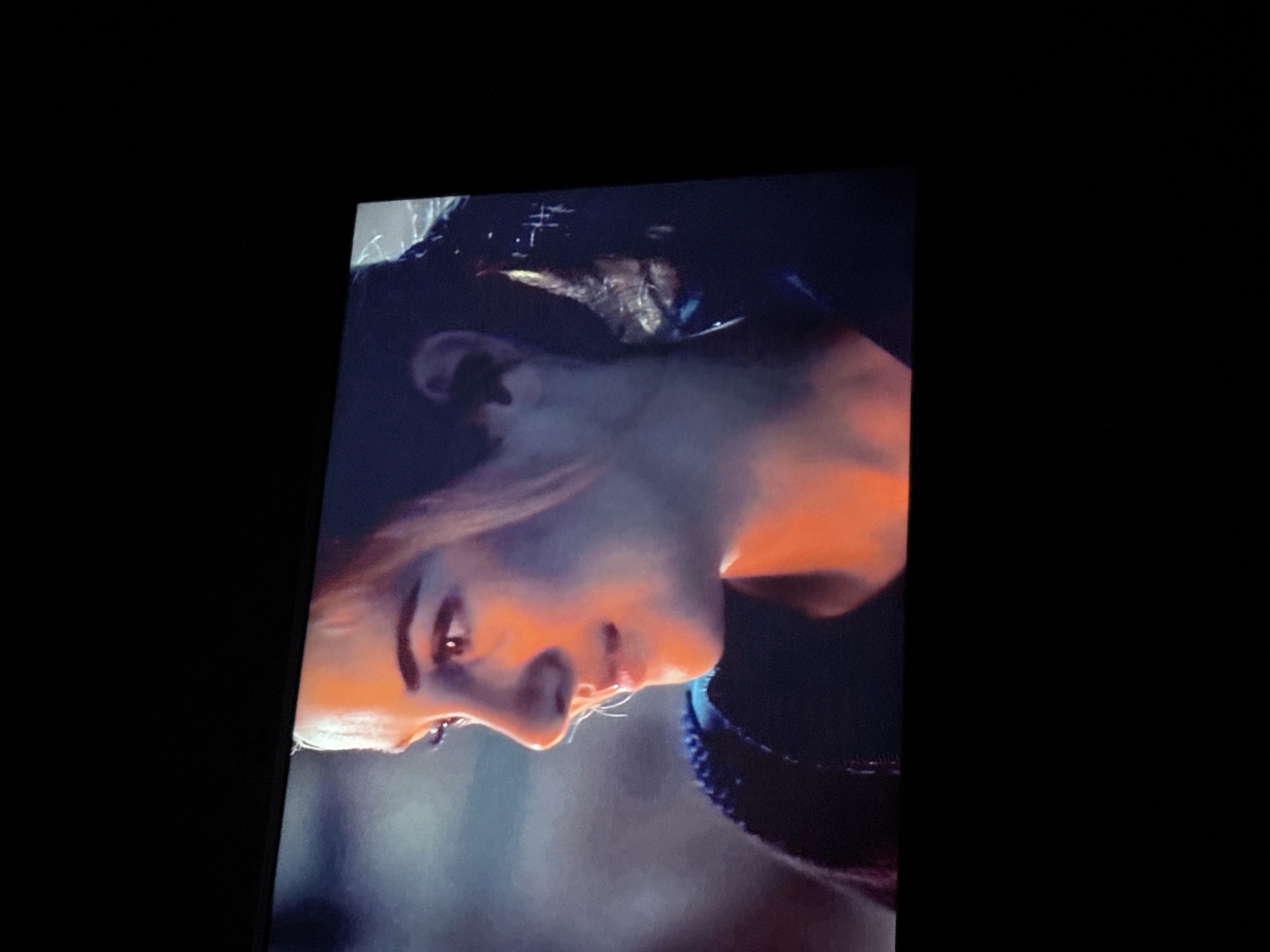 The image shows a close-up of a person's face, tilted to the left. The person has light skin and appears to be illuminated by an orange light source. The background is dark and blurry. The image doesn't contain enough information to identify the location as Mytishchi, Russia, or to provide further details about subjects, objects, actions, or interactions.