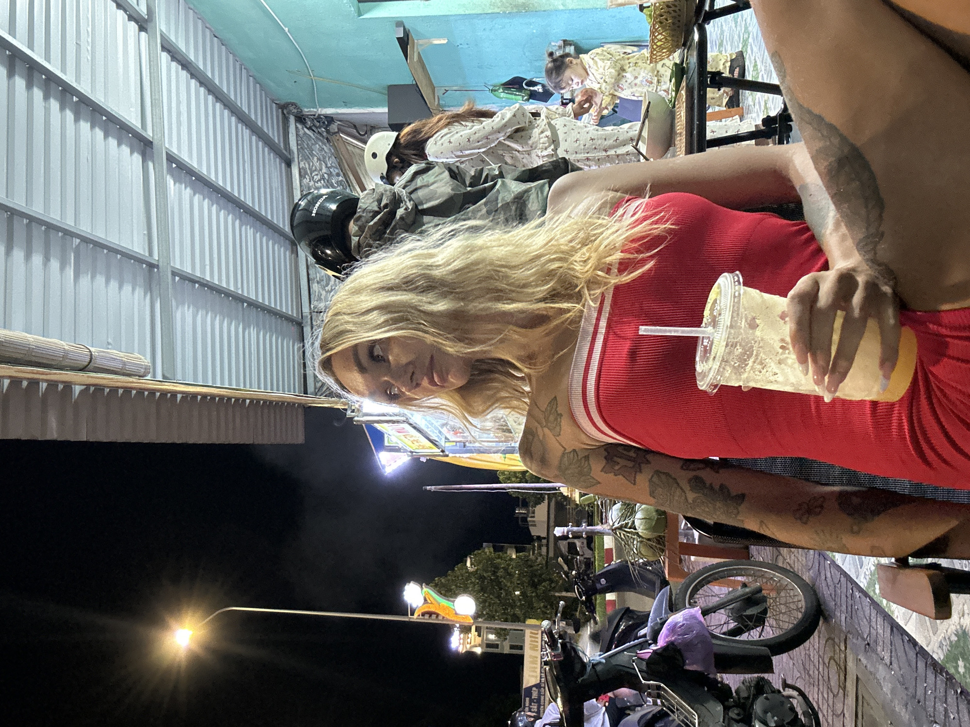 A fair-skinned woman with blonde hair and tattoos on her arms and legs is seated at an outdoor cafe in Phu Thuy Ward, Vietnam. She is wearing a red strapless dress with white trim and holding a plastic cup with a straw containing a yellow beverage. In the background, several people are visible, including a child wearing a light-colored pajamas, and what appears to be a stall or restaurant. Motorbikes are parked on the left side of the image. The scene is lit by a streetlamp and the lights from the cafe, indicating it is nighttime. The overall environment suggests a casual, open-air dining or gathering space typical of street-side establishments.