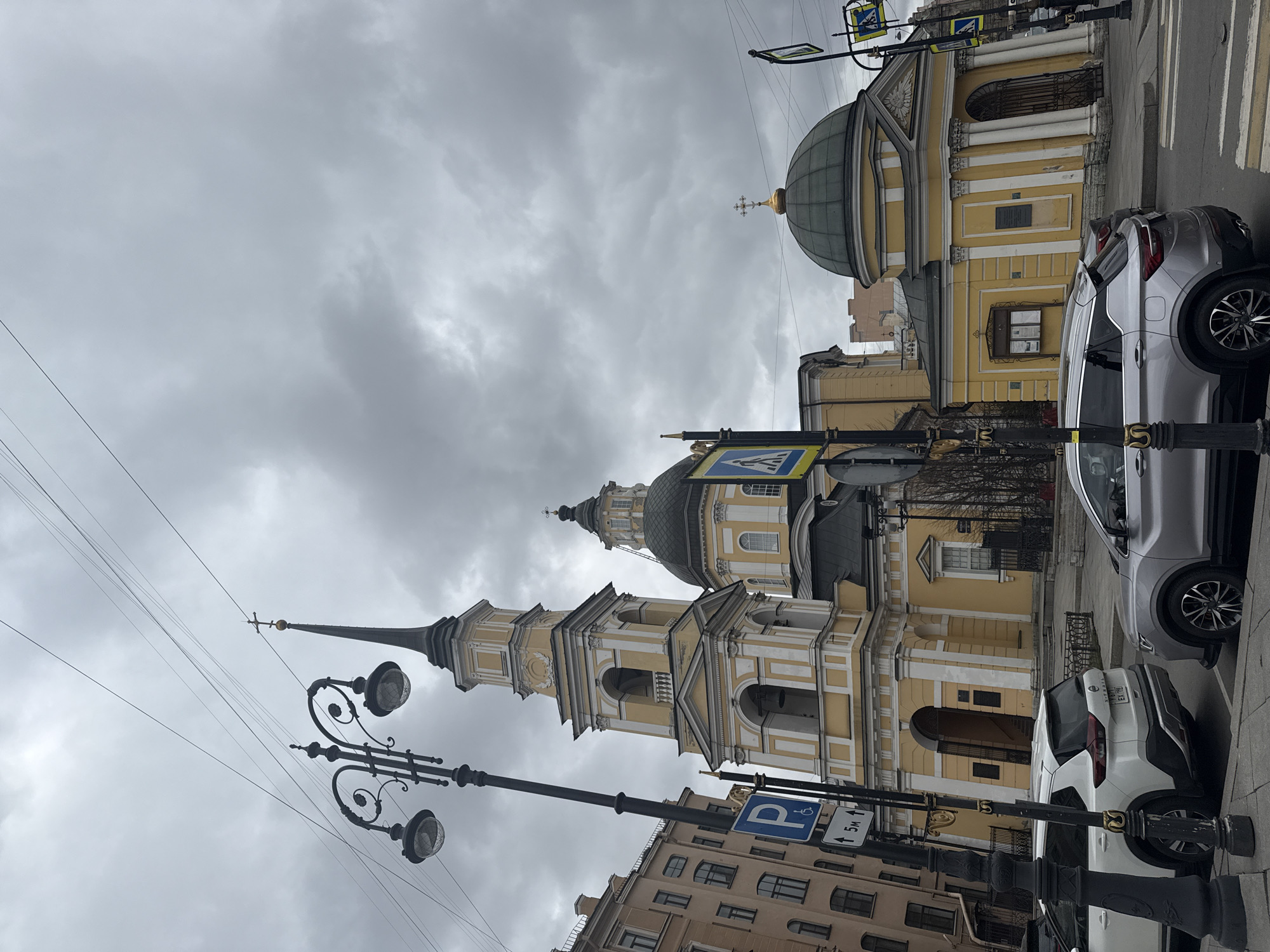The image shows a street scene in Saint Petersburg, Russia, featuring the Church of the Savior on Sennaya Square. The church is characterized by its yellow and white facade, prominent bell tower with a spire, and various domes. The sky is overcast with heavy clouds. Street furniture visible in the scene includes ornate lampposts, pedestrian crossing signs, and parking signs. Two parked cars, one silver and one white, are visible along the side of the street adjacent to the church. The surrounding buildings are visible, suggesting an urban setting with multiple-story buildings. No people are clearly discernible in the image.
