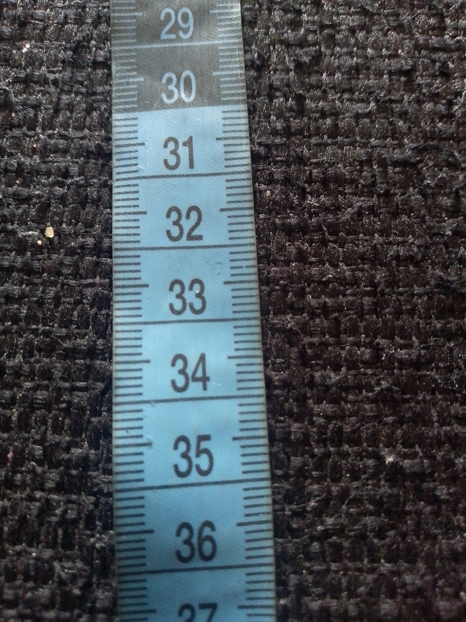 The image shows a close-up of a blue measuring tape with black markings against a textured dark fabric. The measuring tape is marked with numbers indicating units of measurement, with visible numbers including 29, 30, 31, 32, 33, 34, 35, and 36. The fabric appears to be a coarse weave. There are no people or discernible actions captured in the image. The image does not contain any contextual information that would suggest a location in Jauro Yinu, Nigeria, or any other specific geographic area.