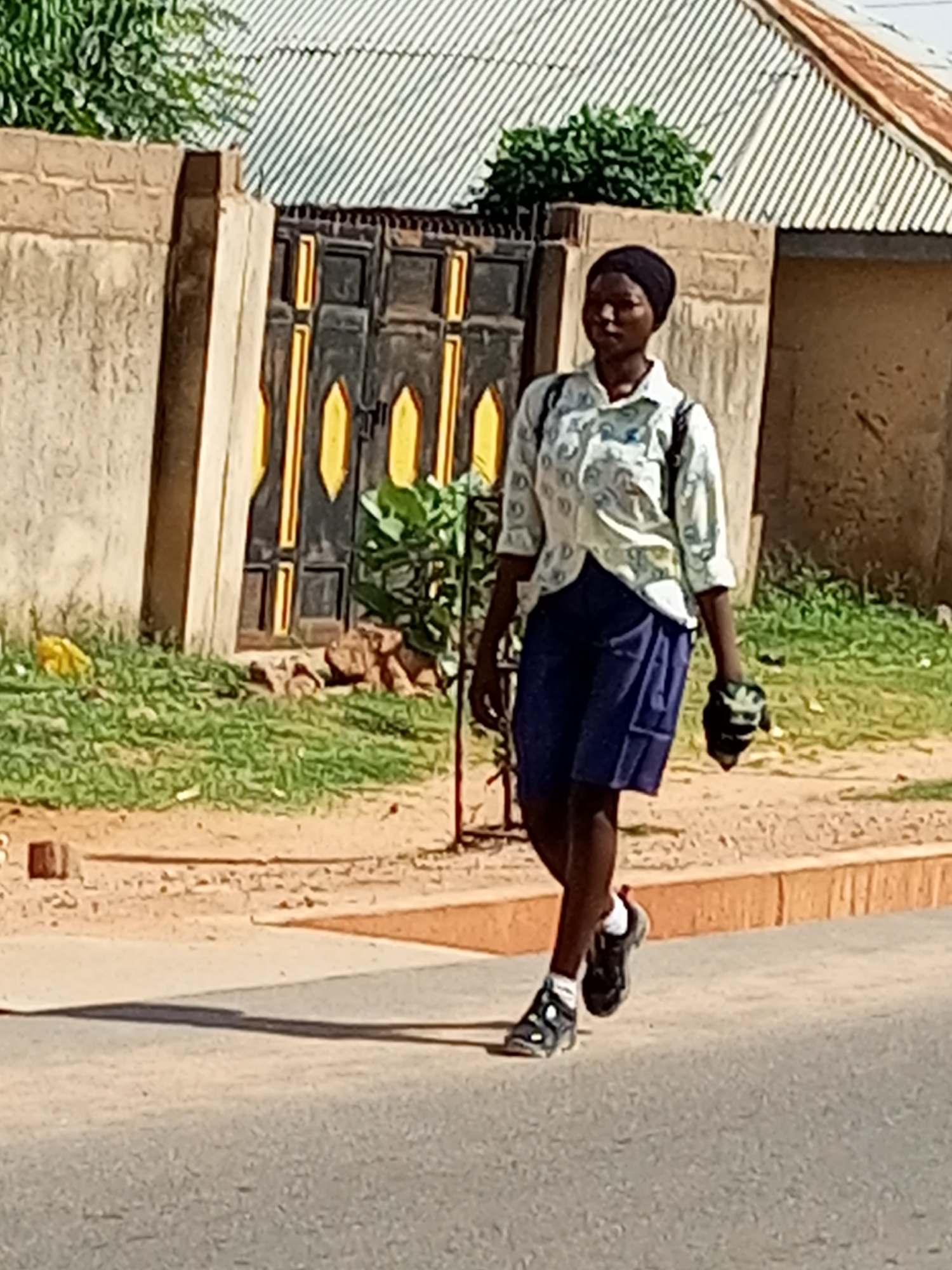 A young person wearing a black headscarf, a light-colored patterned shirt, and a blue skirt walks on a paved road in Gombe, Nigeria. The person is wearing black sneakers with white socks and carrying a black object in their right hand. The background features a tan brick wall with a black and yellow decorative metal gate. Beyond the wall, a building with a corrugated metal roof is visible, along with green foliage. The ground in front of the wall is covered in grass and dirt. The road surface is asphalt.