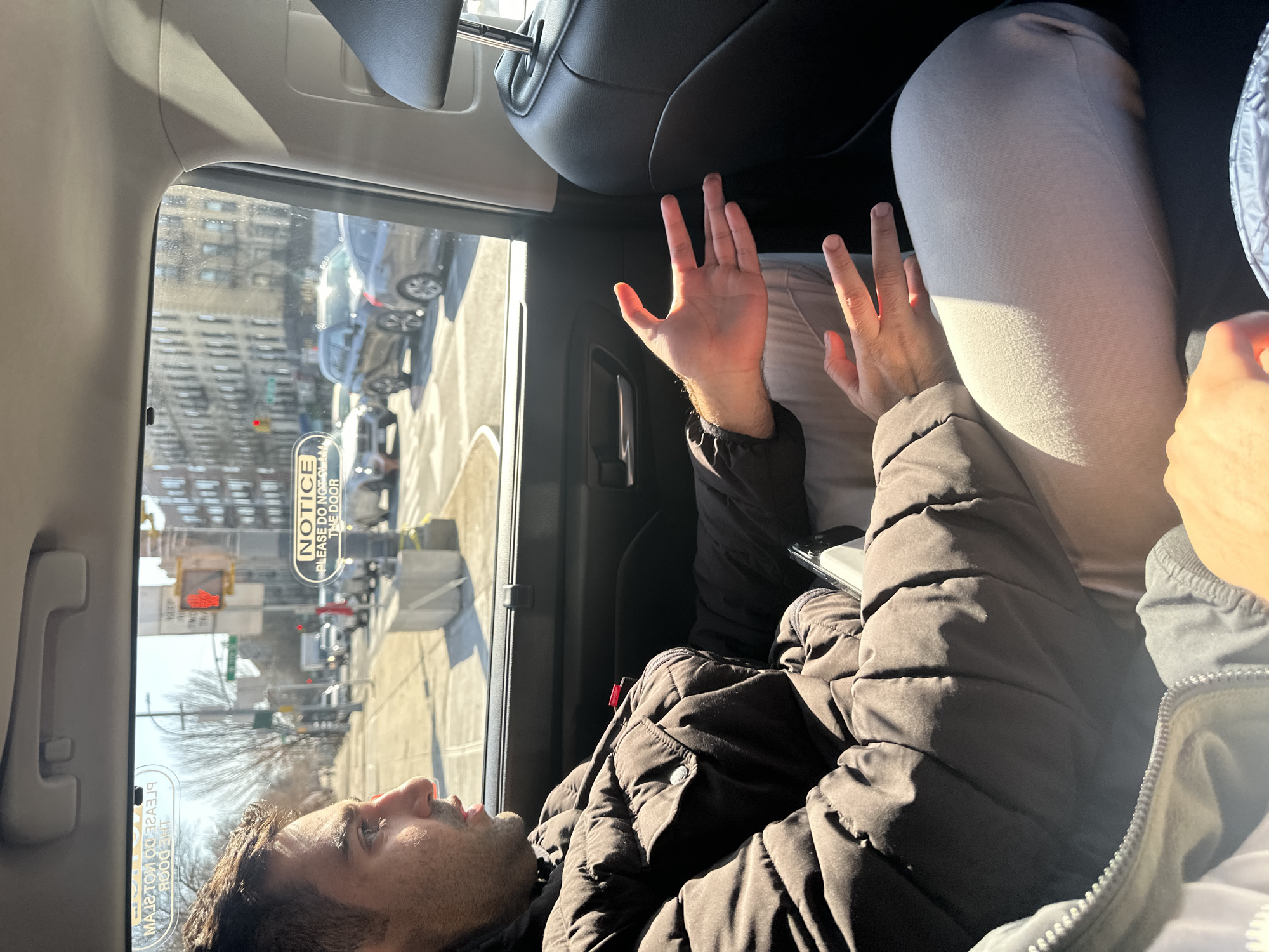 The image depicts a man lying down in the back seat of a car, presumably a taxi or rideshare vehicle, in what appears to be New York City. The man is wearing a dark-colored, puffy winter coat and light-colored pants. His face is visible as he gazes out the window. Both of his hands are raised. The outside scene visible through the window shows city buildings, traffic, and a traffic light. A "NOTICE" sticker is visible on the window.