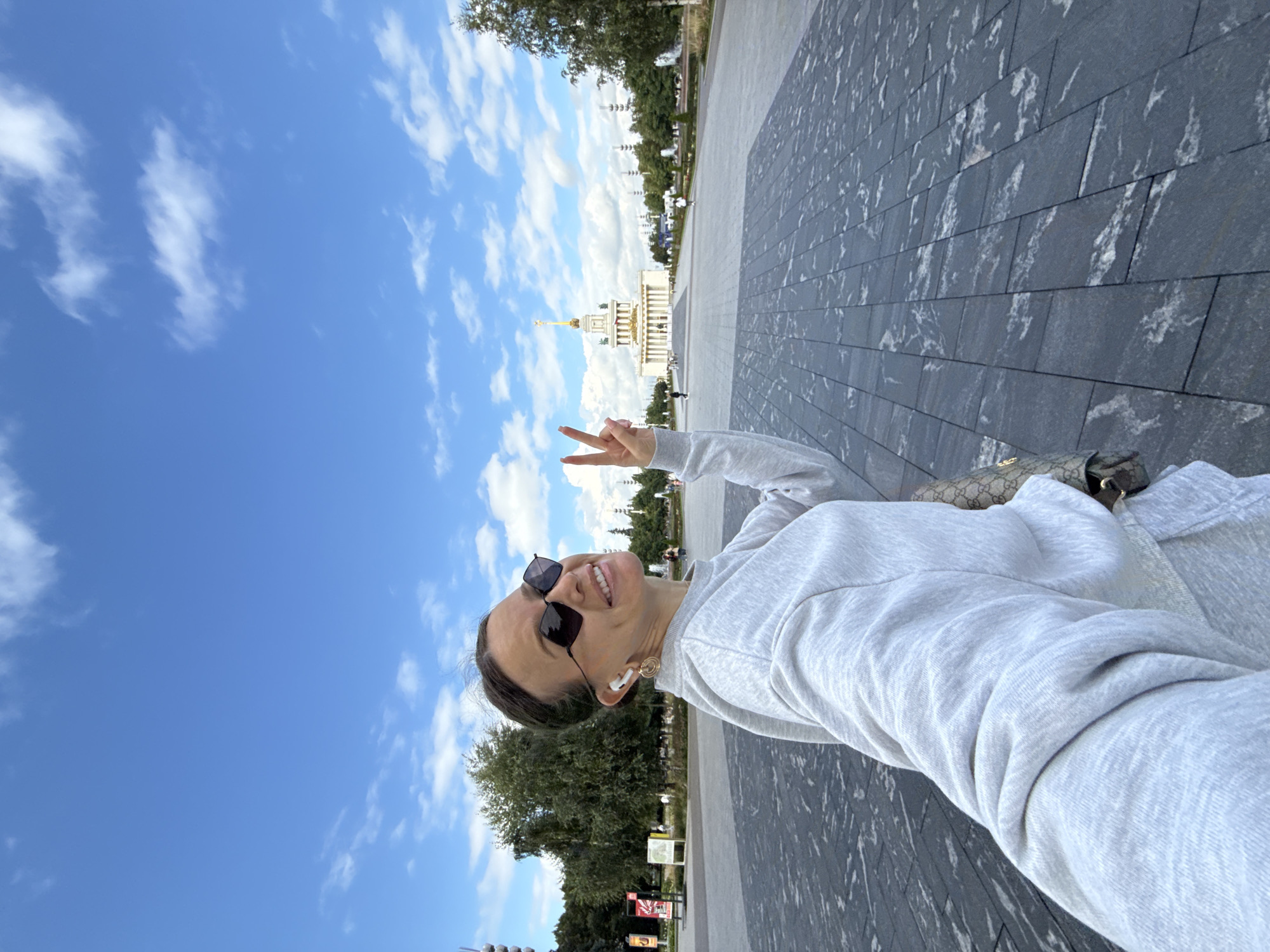 A young woman with dark hair pulled back and sunglasses is taking a selfie outdoors. She is wearing a light grey sweatshirt and is making a peace sign with her right hand. She has earbuds in her ears and a gold earring in her visible ear. She is carrying a designer handbag. The background shows a large, paved plaza with a white building with a golden spire in the distance, possibly representing a landmark. There are trees and other people in the background. The sky is blue with scattered white clouds. The location is identified as the city of Khimki, Russia.
