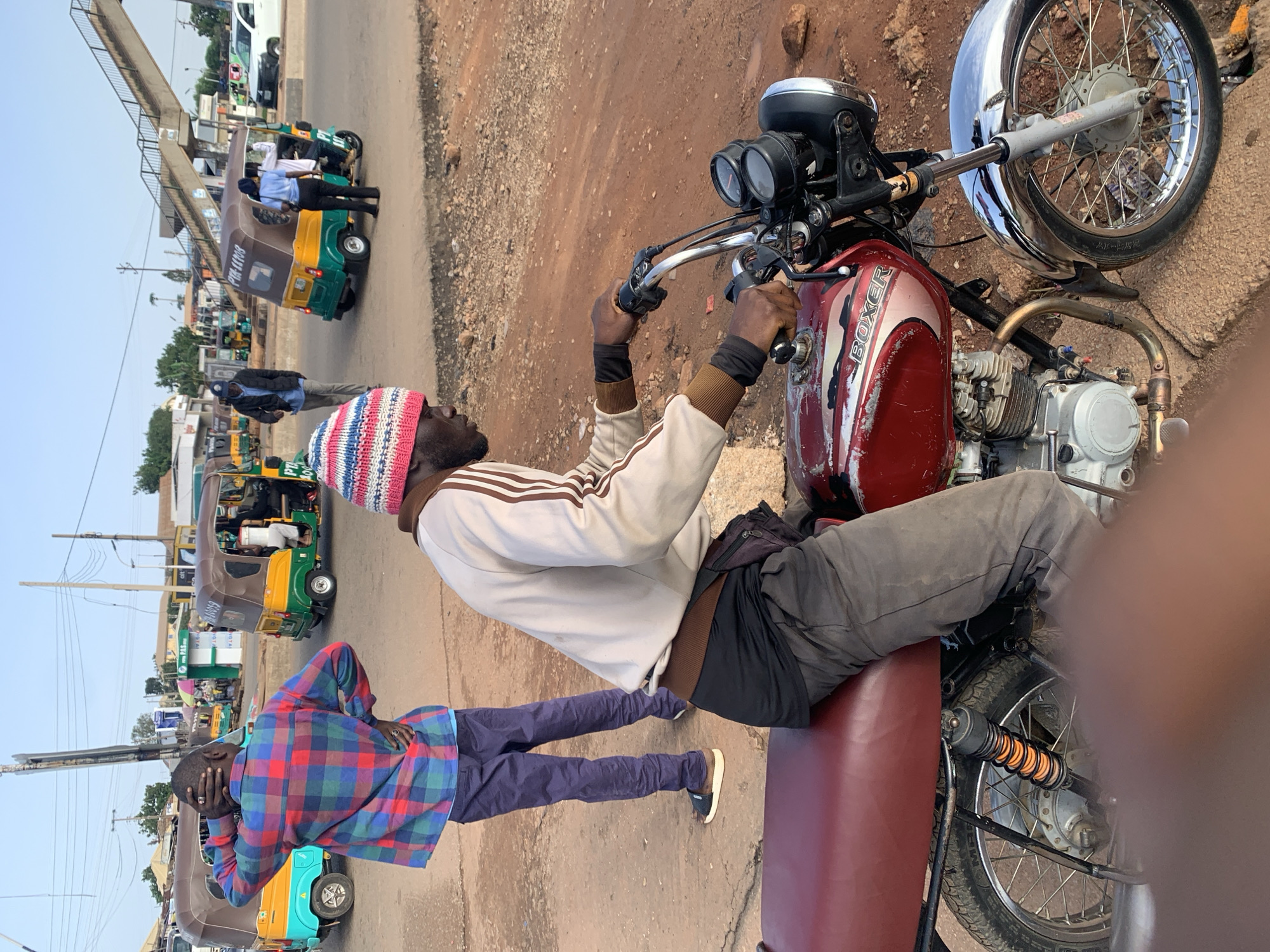This image captures a street scene in Gura Top, Nigeria. Two men are the primary subjects, both appearing to be of African descent. One man is seated on a red motorcycle, wearing a striped hat, a light-colored jacket with brown stripes, and dark pants. His hands are on the handlebars. The second man stands in front of the motorcycle, facing away from the camera. He is wearing a red, blue, and green plaid shirt and dark pants, with his hand on his head.
In the background, several auto-rickshaws, also known as "keke," are visible on a paved road. One of the auto-rickshaws has "PTA LIMA" printed on its side. Another has "PTA Mo" visible. The setting appears to be a busy area, with overhead wires and some foliage suggesting an urban or semi-urban environment. The lighting suggests it is daytime, possibly late afternoon given the angle of the shadows. The overall mood seems to be one of everyday activity and commerce.