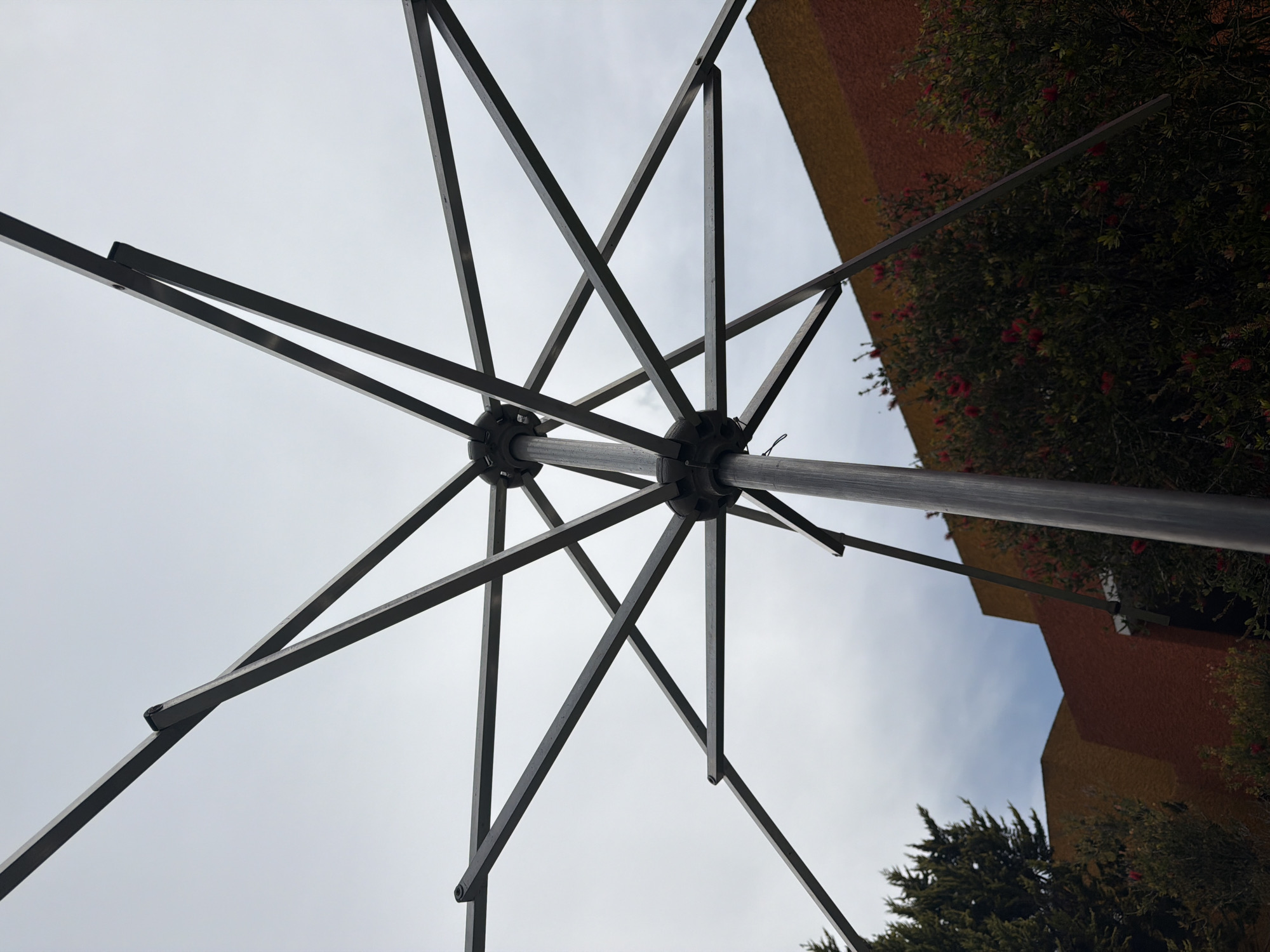 The image shows the underside of an open umbrella, presumably a patio umbrella, set against a background of a partly cloudy sky, trees, and the side of a building. The umbrella frame is made of metal and features a central pole extending upwards from the perspective. The frame consists of multiple supports radiating from two central hubs.
The top portion of a building, possibly two-toned (orange and yellow), is visible, along with greenery suggesting trees and bushes. The scene appears to be outdoors, likely in an urban setting due to the presence of the building, though the specific location within the city of Unknown, Unknown, is not discernible from the image alone. There are no people visible in the image.