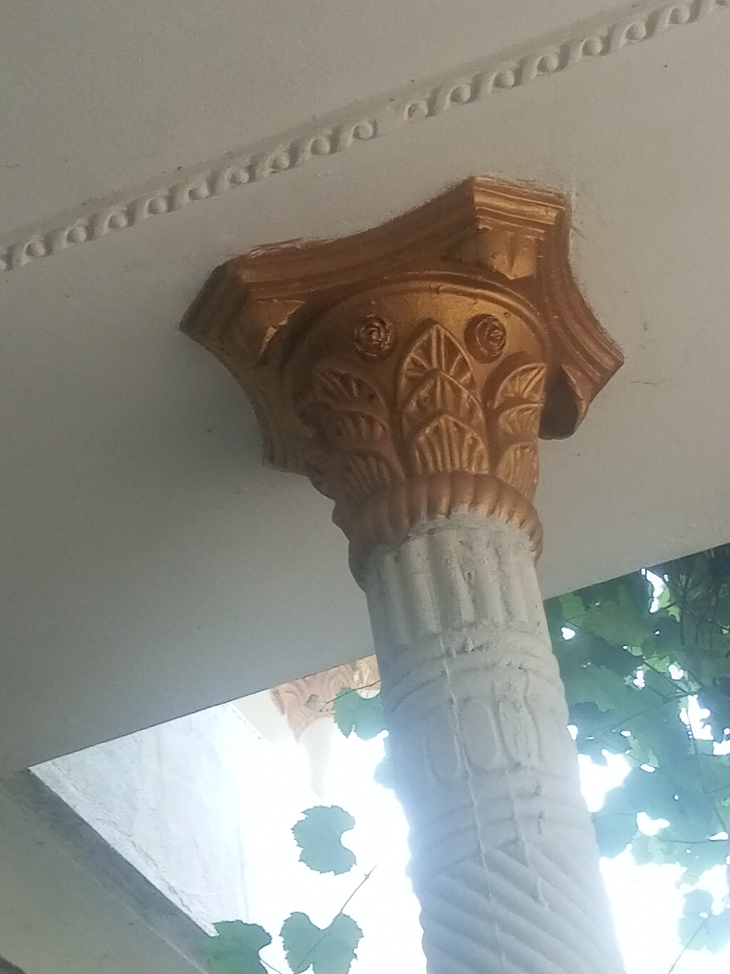 A close-up, angled shot focuses on the ornate capital of a white, fluted column, adorned with a golden, decorative element featuring stylized leaf and rose motifs. Above the capital, a white ceiling exhibits a decorative border of interlocking semi-circular moldings. In the background, lush green foliage suggests an outdoor setting, with a bright light source behind the leaves, possibly indicating sunlight filtering through. The image does not provide any information about a specific location in Mansehra, Pakistan, nor does it depict any people or actions.