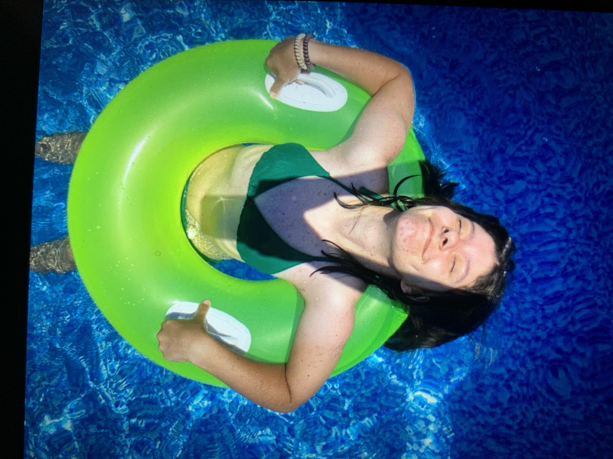 A person is floating in a swimming pool with a bright green inflatable ring. The person is wearing a dark green bikini and has dark hair. Their face is turned upwards, eyes closed, with a relaxed expression. Their arms are resting on the ring, and they are holding onto white handles. The water is a vibrant blue, with the rippling surface creating patterns. Feet are visible submerged in the water. The scene suggests a leisure activity in a pool. There is no information in the image to indicate a location in Lymanka, Ukraine.