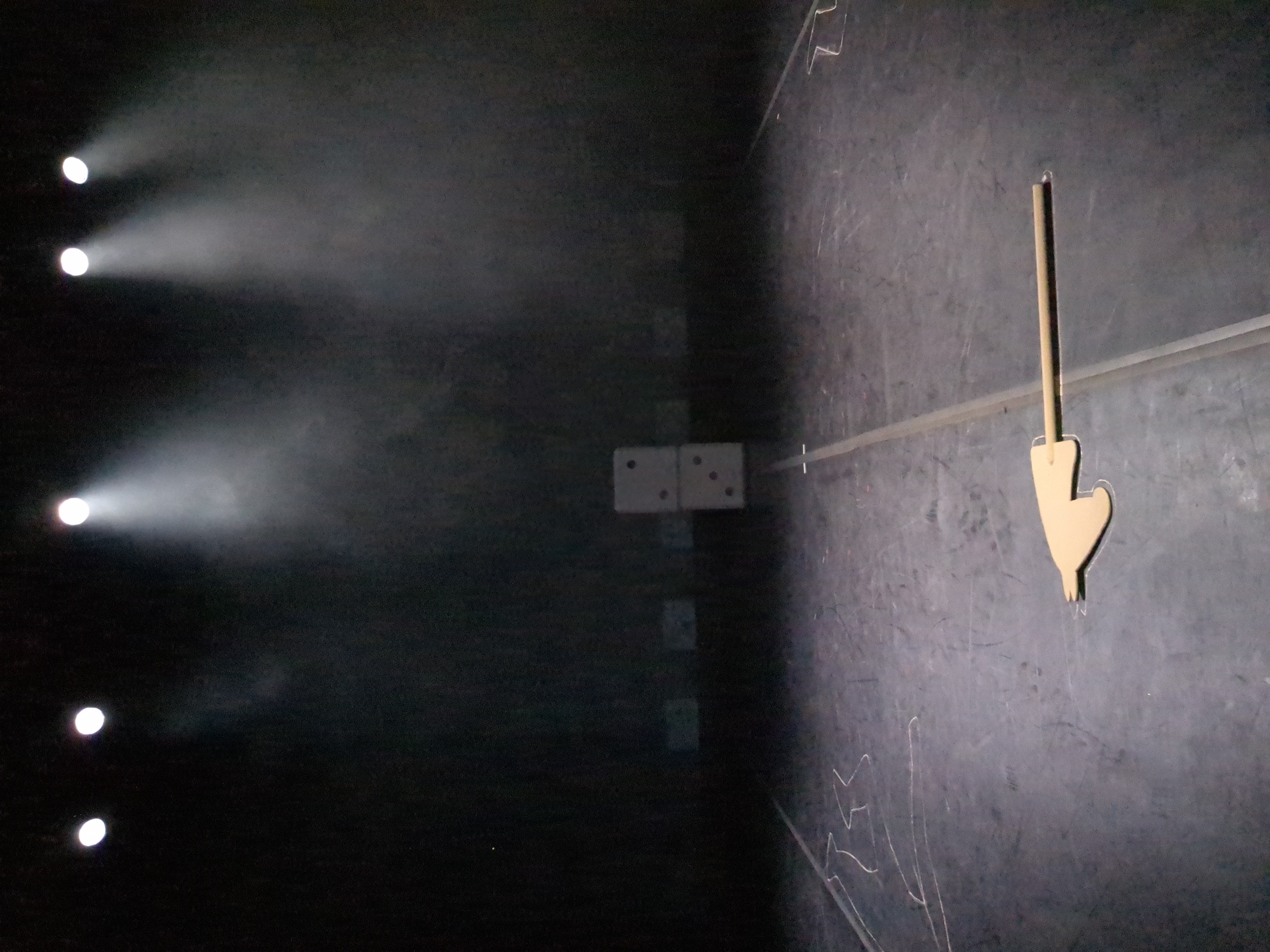 The image shows the interior of a dark space, possibly a room or stage, with an unevenly illuminated black wall on the right side and a dark area on the left punctuated by several bright spotlights. The right wall has a light-colored, vertical, hand-shaped object hanging from it, and chalk markings appear near the bottom. Further back, closer to the center of the frame, are two lighter-colored rectangular shapes with dots, possibly dominoes or some other similar gaming pieces, affixed to the wall. The scene lacks clear indications of a specific location within Saint Petersburg, Russia, and does not show any people or distinct interactions. The dominant features are the lighting contrast and the objects attached to the right wall.