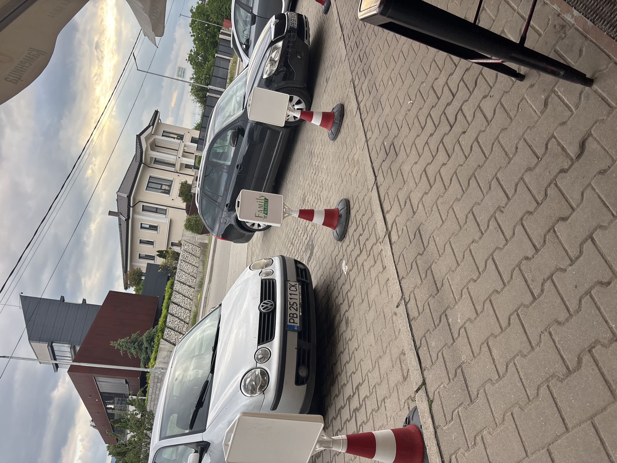 The image depicts a street scene in Plovdiv, Bulgaria. Two cars, a white Volkswagen Polo with license plate "PB 2511 CK" and a dark-colored Skoda Octavia with license plate "PB 5500CE," are parked along a paved sidewalk. Red and white traffic cones with white square signs are positioned around the cars. The signs on the cones appear to be advertisement. Buildings are visible in the background. One building is a modern structure with a dark gray top and a rust-colored section. The sky is overcast with a mix of clouds and sunlight. There are no people visible in the frame.
