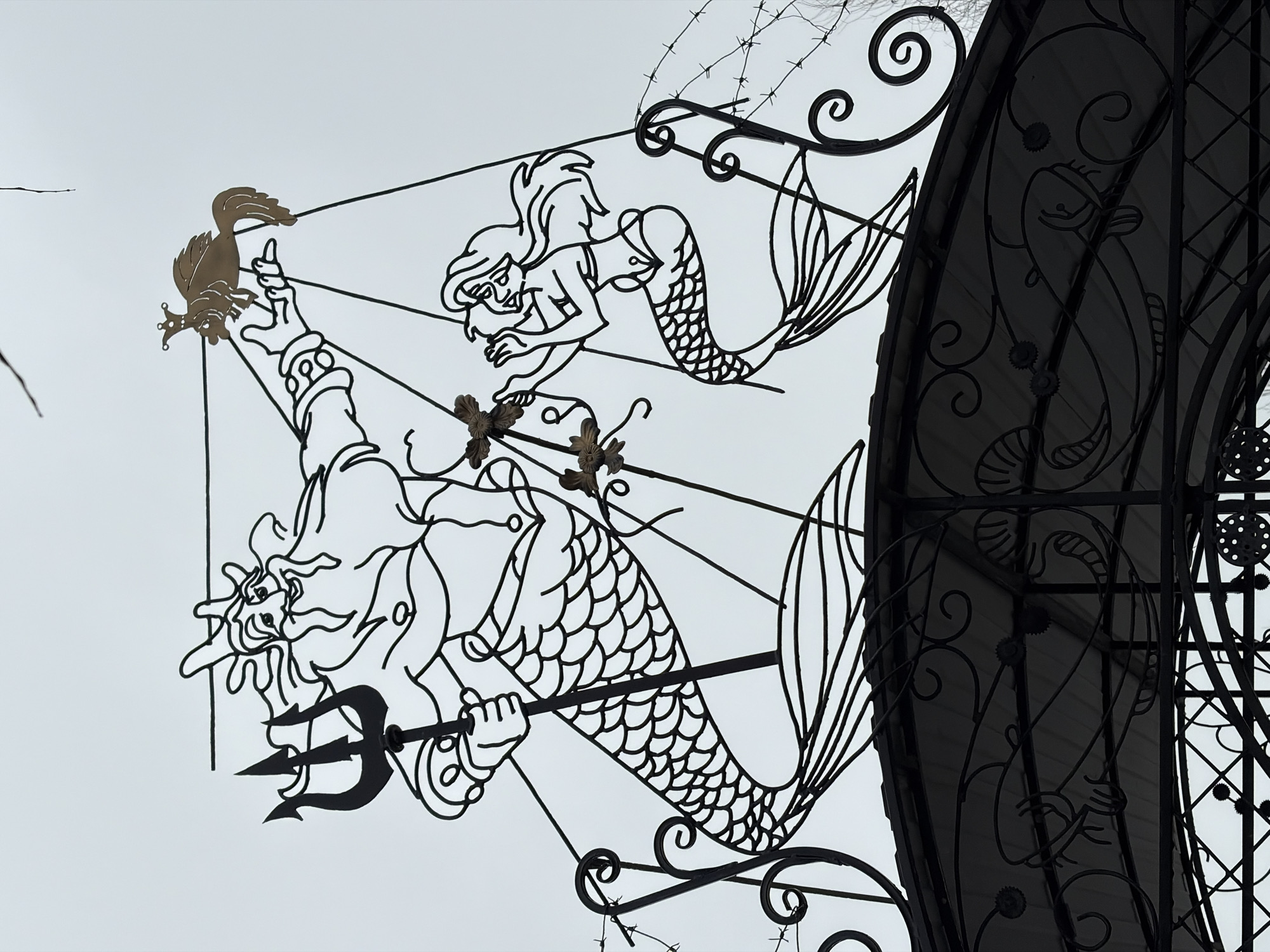 The image shows an ornate metal artwork, possibly part of a gate or fence. It depicts two main figures: Poseidon/Neptune with a trident and a mermaid. Above them is a smaller mermaid and atop Poseidon's extended arm is a bird-like creature with a crown. The background is a cloudy sky. To the right is a partially obscured structure with wrought iron designs, including depictions of fish, suggesting an aquatic theme. The location is Chaikovsky, Russia.