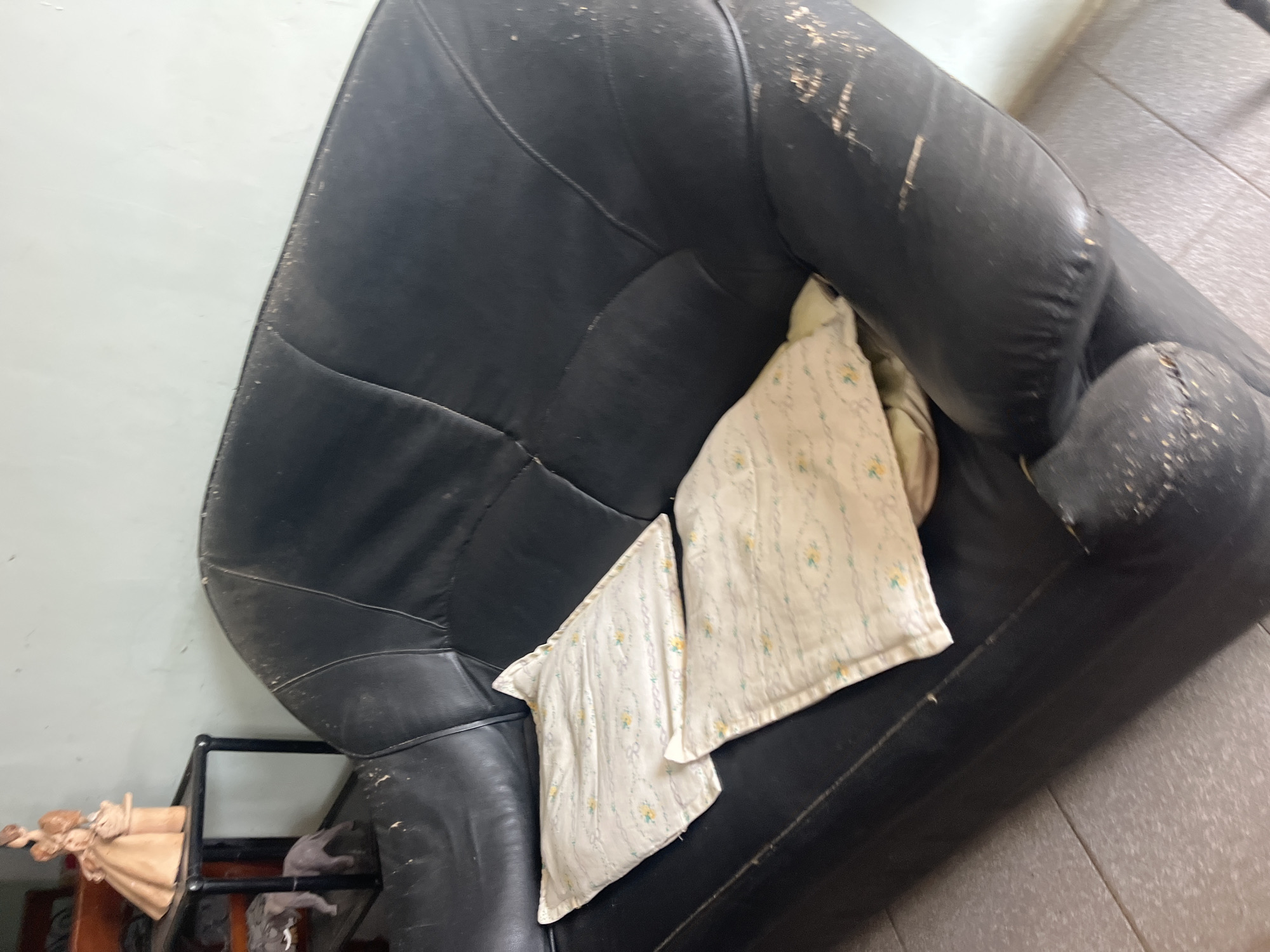 This image captures an indoor scene in Santa Ana, Venezuela, centered on a black, two-seater sofa. The sofa's leather-like upholstery is significantly deteriorated, displaying extensive peeling, flaking, and abrasions across its arms, seat cushions, and backrest, exposing a lighter substrate material. Two light-colored, square pillows, adorned with a pattern of small yellow flowers and green vines, rest on the sofa's seat. Adjacent to the left side of the sofa stands a black, two-tiered end table. The top tier supports a beige figurine of a woman holding a basket. The lower tier holds two grey elephant figurines, varying in size. The floor consists of light grey square tiles. The background features a plain, light-colored wall. No persons are depicted, and no active events or interactions are observed.