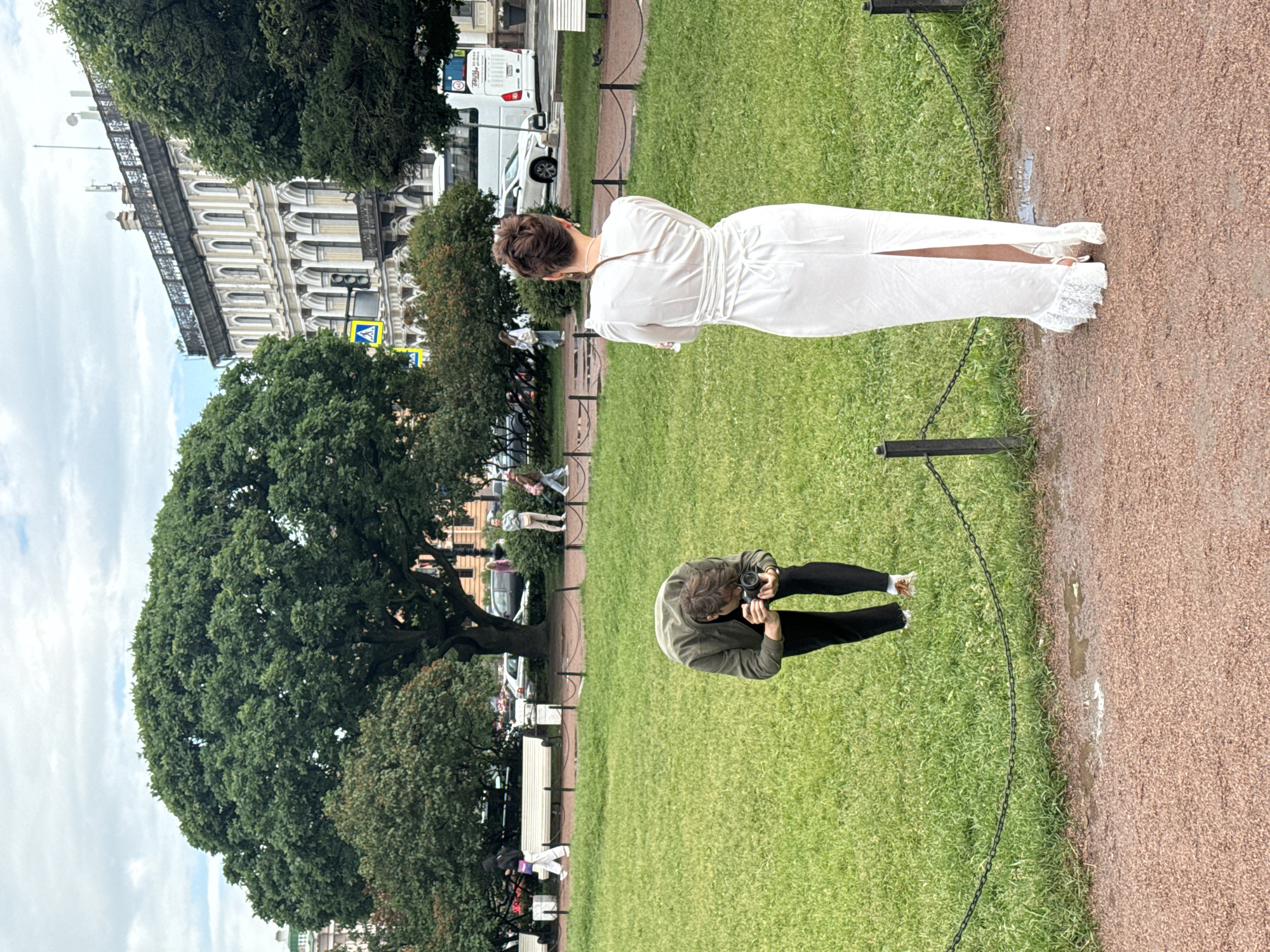 In Saint Petersburg, Russia, on a cloudy day, a photographer in a green jacket and black pants is crouched on the grass, taking a picture with a camera. A woman in a long white dress with lace trim and high heels stands facing away from the camera, posing in front of a black chain fence. The background features a large tree, a building with ornate architecture, and other people walking on a path. The ground is a mix of grass and gravel.