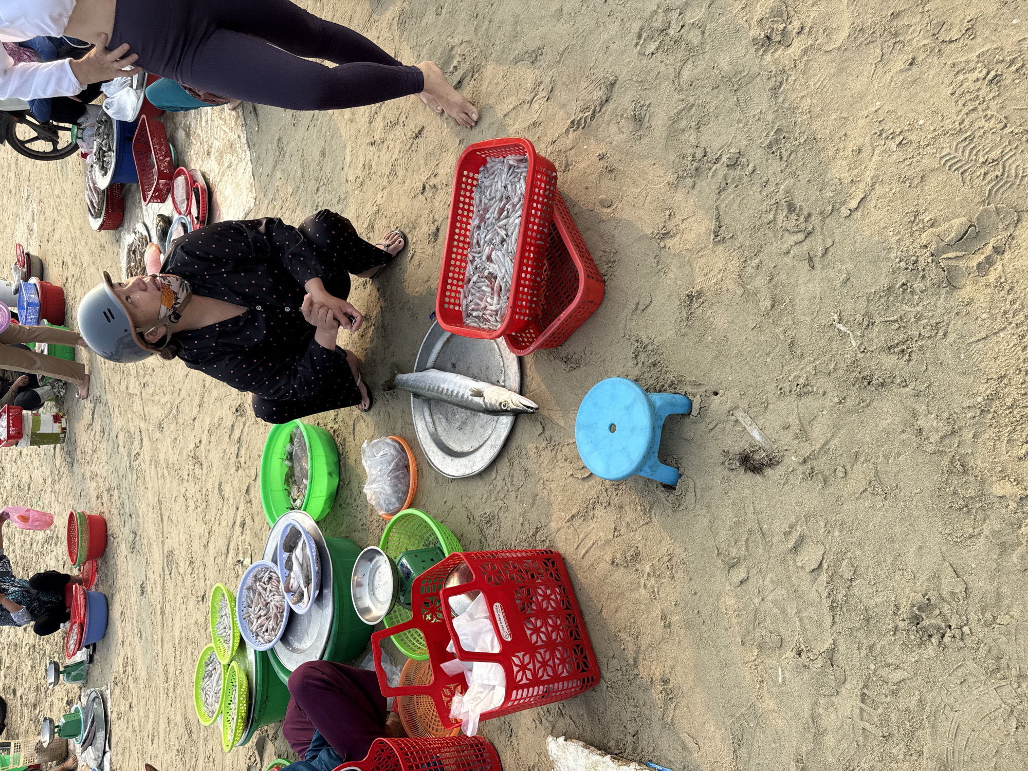 The scene depicts a sandy beach in Đà Nẵng, Vietnam, where several people are selling fish. A woman wearing a black patterned outfit and a helmet is seated amongst baskets and metal trays filled with various small fish. She is interacting with someone out of frame. Another person, wearing leggings, stands nearby. Red and green plastic baskets and metal trays containing fish are scattered on the sand. A small blue stool sits to the right of the seated woman. A motorcycle is partially visible in the background. The scene suggests a local seafood market on the beach.