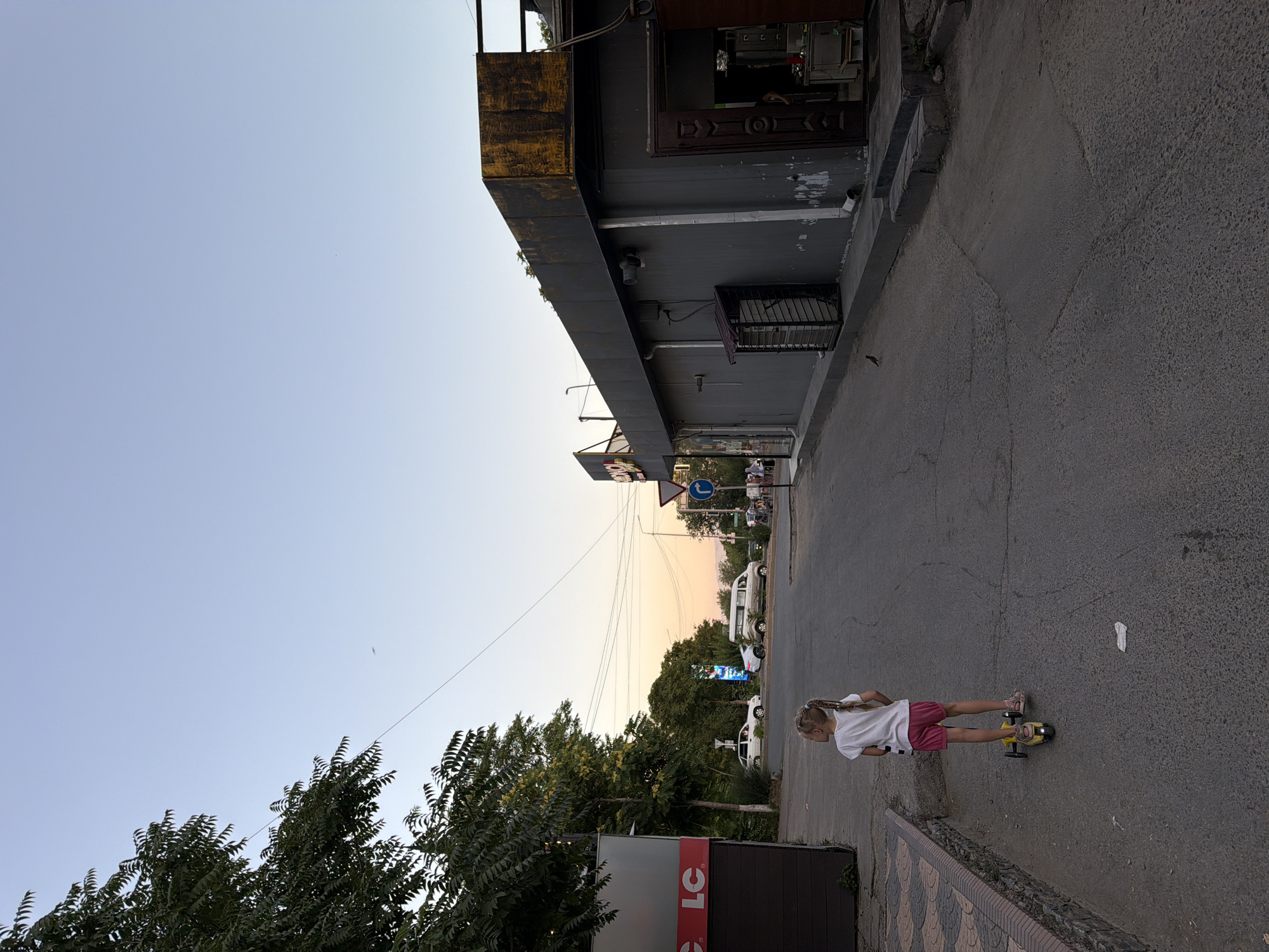 In Shymkent, Kazakhstan, a young girl with blonde hair braided down her back is riding a yellow skateboard on an asphalt road. She is wearing a white t-shirt and pink shorts. The scene is set outdoors, with a building on the left side of the frame and a street stretching into the distance. The building appears to be a commercial establishment with a grey facade and some visible signage, though the text is not clearly legible in the full image. A portion of the building features a distressed yellow facade above a dark grey overhang.
The sky is clear and a pale blue, suggesting it is either early morning or late afternoon. The lighting indicates a warm, soft glow, possibly from the setting or rising sun, painting the distant sky with subtle hues of orange and pink. Trees with lush green foliage line the background, and a few cars are visible further down the street. One of the cars is a white van, and another is a white sedan. There are also power lines strung across the sky. The overall atmosphere is relaxed and casual, with the girl enjoying a leisurely activity on the street. A sign with "LC" in red letters is visible on a dark panel near the bottom left of the image, likely indicating a storefront.