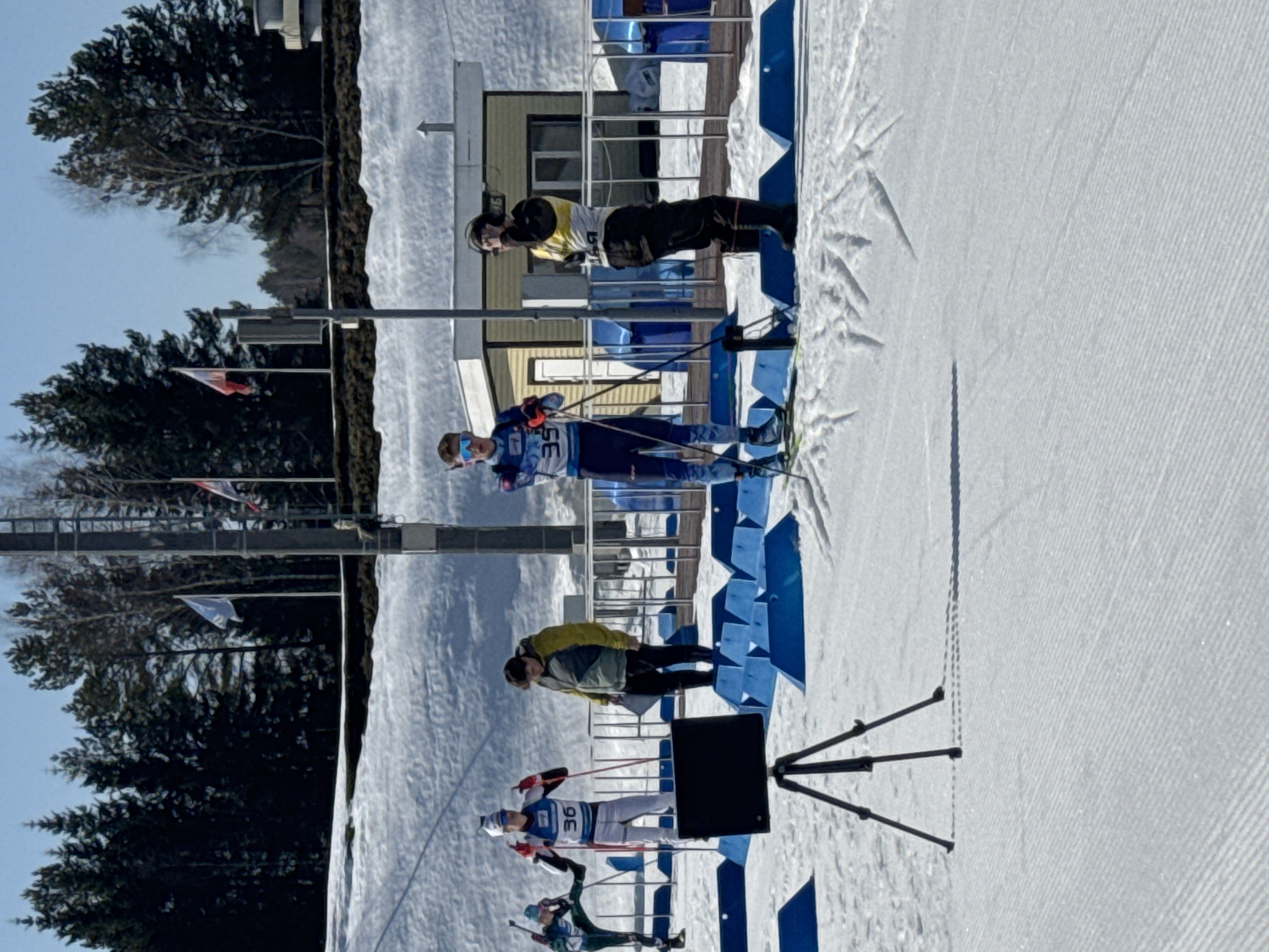 The image displays an outdoor winter sports environment in Прикамский, Russia, under a clear blue sky. The ground is uniformly covered in groomed snow, exhibiting textured patterns from snow-grooming equipment and distinct ski tracks.
In the foreground, a black electronic display screen is mounted on a black tripod, positioned centrally. Multiple blue, triangular-prism-shaped markers are arranged in rows across the snow, likely delineating lanes or boundaries.
Five individuals are visible in the scene:
1. On the far left, partially obscured, a person in a green and blue athletic suit is in an active skiing or stretching posture, with one ski elevated.
2. To the right, a person in a white and blue ski suit, marked with bib number "36," stands with ski poles, appearing to gesture with gloved hands.
3. Behind the blue markers and to the right of person 36, an individual in a dark jacket with yellow and green accents stands holding a clipboard or documents.
4. Further right, a person in a blue and white ski suit, wearing bib number "35" and blue-mirrored sunglasses, stands on skis, holding ski poles.
5. On the far right, near a building, an individual in dark clothing with a yellow bib is standing, seemingly adjusting headphones.
In the mid-ground, a single-story, light-colored building with a dark roof is present. To its right, another building with a rounded roof structure is partially visible. Several vertical poles are positioned in the background, one of which displays a light-colored flag, while another carries the Russian flag (white, blue, red horizontal stripes). A dense treeline of dark conifers and lighter deciduous trees forms the distant background, with snow-covered inclines visible below the trees. Strong shadows are cast on the snow, indicating direct sunlight.