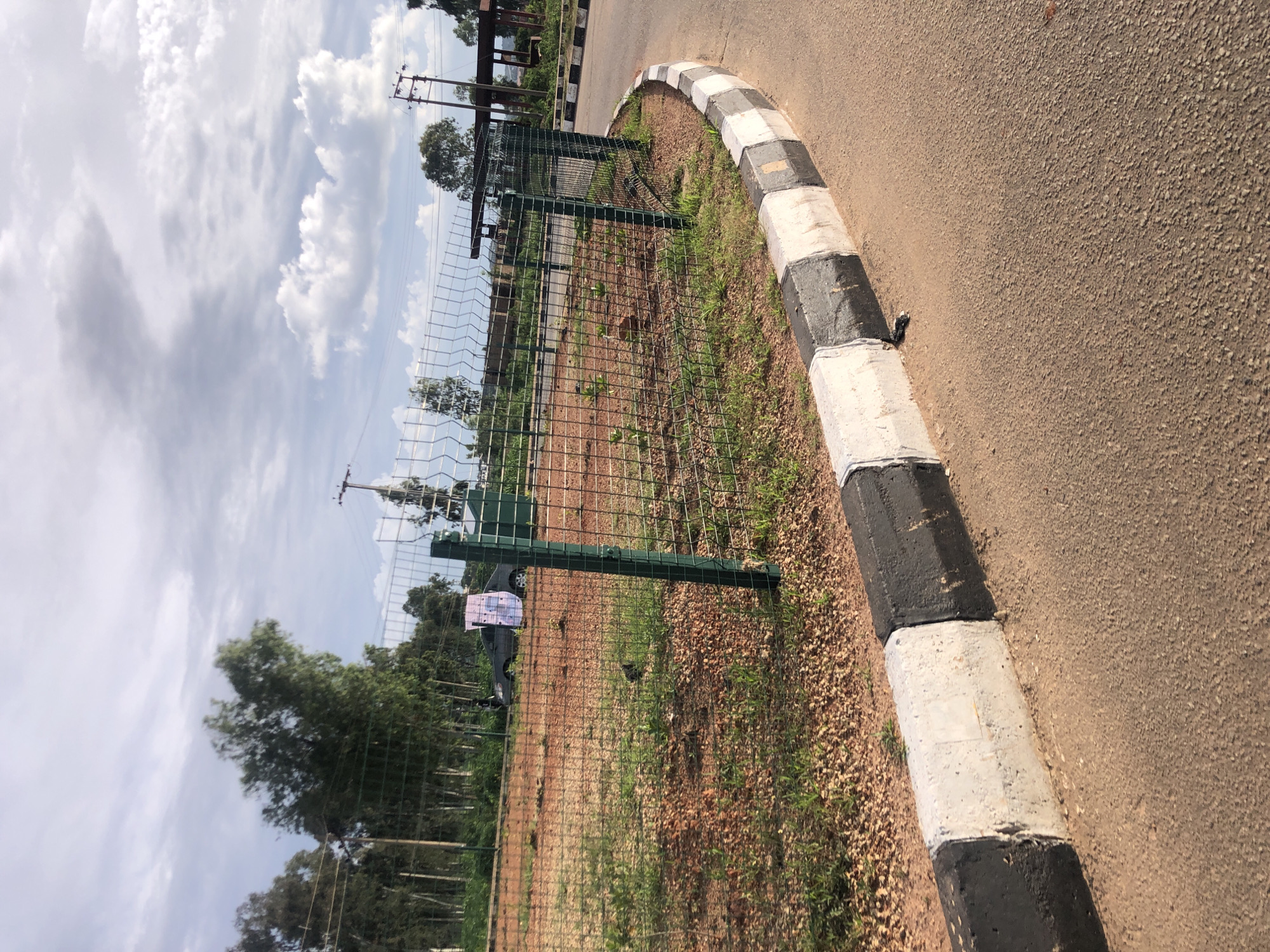 The image depicts a roadside scene in Jos, Nigeria. A paved road with a distinct black and white striped curb runs along the right side of the frame. To the left of the curb, a wire mesh fence encloses an area with sparse vegetation and reddish-brown earth. Beyond the fence, several trees are visible, some with dense foliage, and in the distance, there appear to be utility poles and possibly some buildings.
The sky is partially obscured by clouds, suggesting it is daytime. The lighting indicates it's likely morning or afternoon. There is no discernible activity or event taking place; it appears to be a static scene. A small sign with some text is affixed to one of the fence posts, but the text is not legible. The overall impression is of an urban or semi-urban environment, possibly an undeveloped plot of land next to a road.