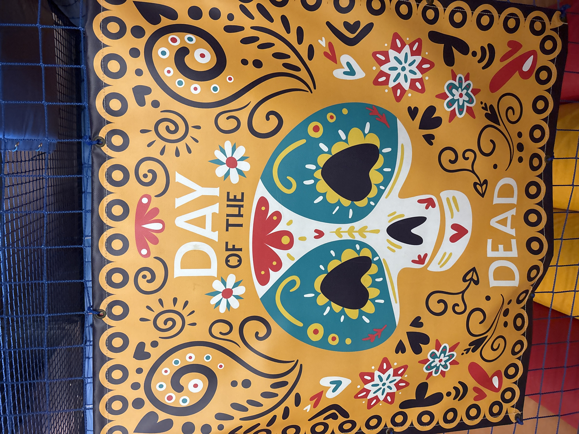 The image shows a colorful banner with the words "DAY OF THE DEAD" displayed prominently. The banner features a stylized skull adorned with floral and swirly patterns in shades of orange, yellow, teal, red, white, and black. The banner is hung outdoors, with a blue net and dark blue fabric visible in the background, suggesting it might be part of a temporary event or decoration. The lighting indicates it is daytime. There is no visible indication of the location being Dubai, United Arab Emirates.