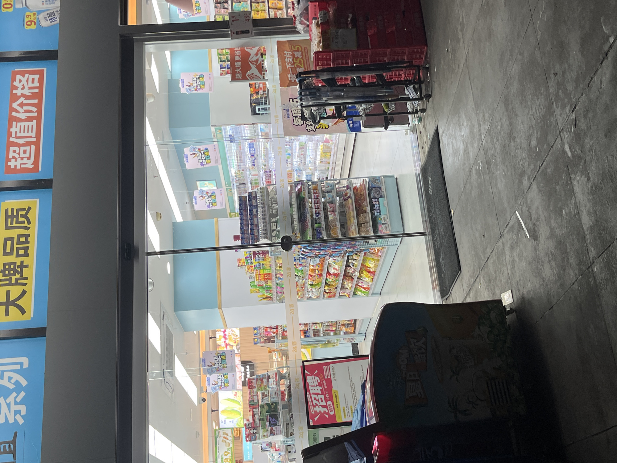 This is a medium shot, exterior view of a convenience store entrance in Yuzhong District, China. The automatic glass doors are open, revealing shelves stocked with various merchandise. Above the doors, bright blue and yellow signage with Chinese characters is visible. To the left of the entrance, a red and black vending machine with colorful graphics is positioned on the sidewalk. The pavement in front of the store is made of grey concrete slabs. Inside the store, aisles of products are illuminated by fluorescent lighting, and more signage with promotional offers is displayed. There are no people immediately visible in the doorway or directly in front of the store. The overall scene suggests a retail environment, possibly at dusk or with artificial lighting predominating.