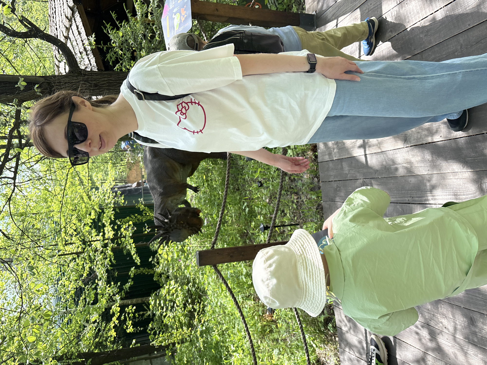 The image depicts a sunny outdoor scene, likely in a park or zoological garden. A woman, wearing sunglasses, a white t-shirt with a Hello Kitty design, and blue jeans, stands on a wooden platform. Next to her, a young child wearing a light green outfit and a white bucket hat appears to be looking at something in the direction of a dinosaur sculpture. In the background, there's a second adult with their back facing the camera, dressed in khaki pants and a blue shirt. The location includes wooden structures and dense green vegetation, suggesting a natural or simulated natural environment with ropes creating barriers, possibly for a viewing area. The photograph was taken in Izhevsk, Russia.