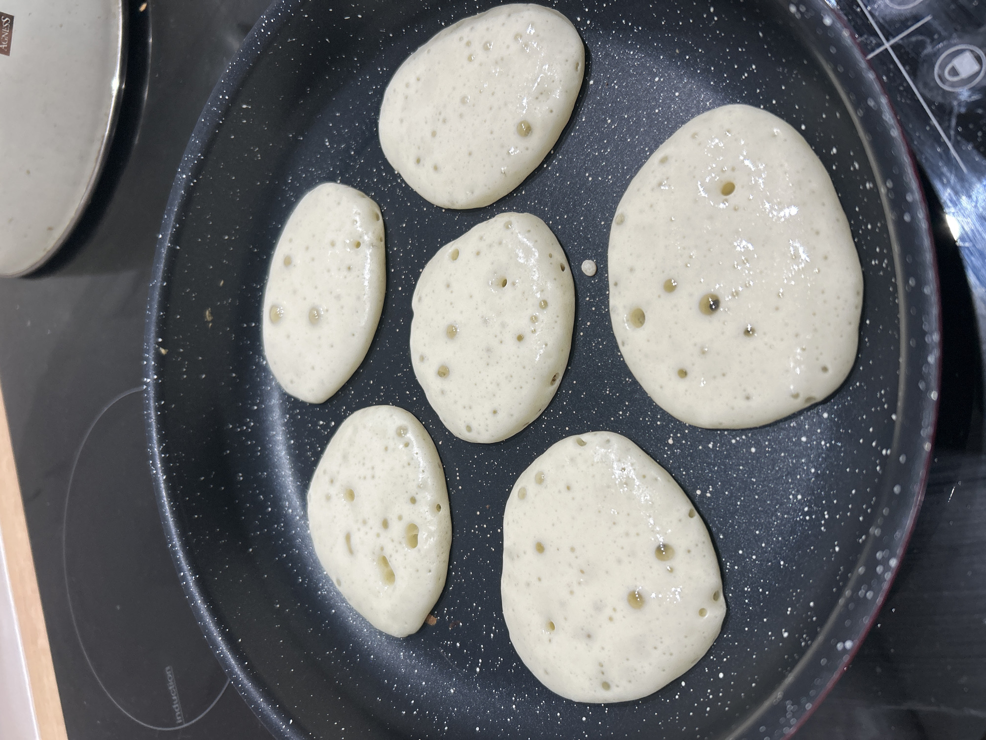 Six irregularly shaped pancakes, likely made from batter, are cooking in a dark, speckled non-stick frying pan. The pancakes are light in color, almost white or pale yellow, and show numerous small bubbles forming on their surface, indicating they are actively cooking. Six pancakes are visible, arranged somewhat haphazardly in the pan.
The pan rests on a black induction cooktop, part of which is visible, particularly to the left and bottom of the pan. A circular marking and the word "induction" can be seen on the cooktop surface to the left. To the upper left, a portion of a shiny metallic pot or lid is visible, with the brand name "AGNESS" partially legible on its side. The scene suggests a home kitchen environment. The overall impression is that food preparation, specifically breakfast or a snack, is underway. There are no people visible, and the time of day is not explicitly indicated but could be morning.