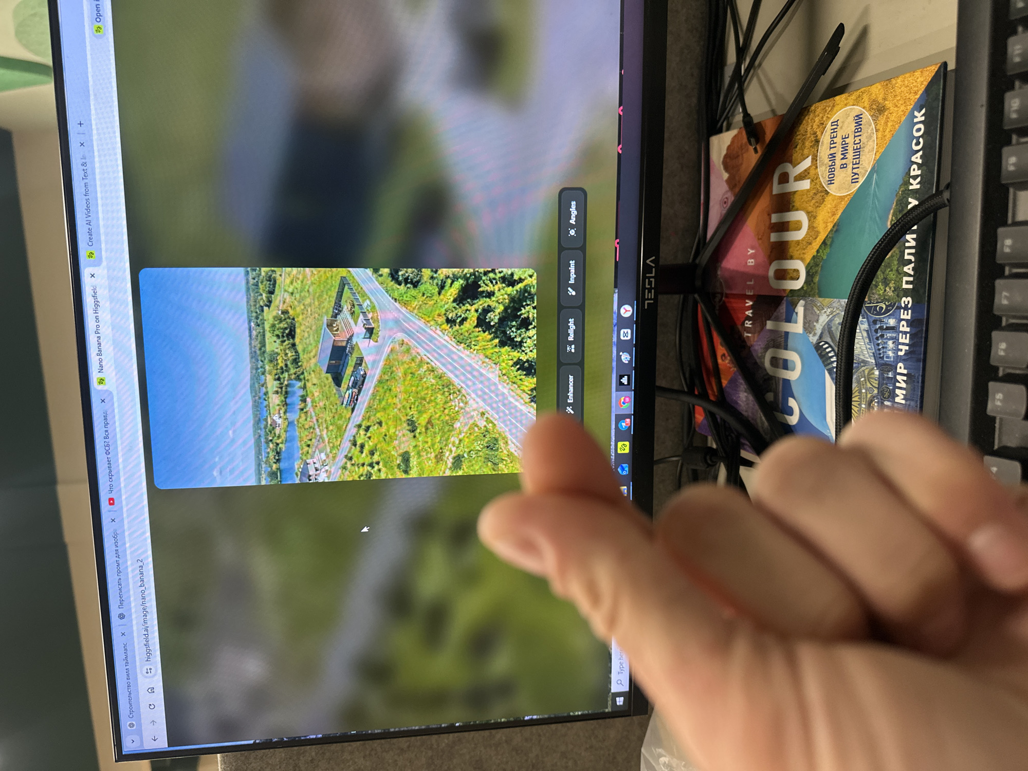 A human hand, out of focus, makes a "finger heart" gesture in the foreground, partially obscuring the lower-left section of a workspace. The primary focus is a black TESLA-branded computer monitor displaying a web browser.
The browser shows multiple tabs open at the top, including titles in Russian such as "Строительство вилл таймлапс" (Construction of villas timelapse), "Переписать промпт для изображ" (Rewrite prompt for images), and "Что скрывает ФСБ? Вся правд" (What does the FSB hide? All the truth), alongside English tabs like "Nano Banana Pro on Higgsfield" and "Create AI Videos from Text & In". The active tab displays the URL `higgsfield.ai/image/nano_banana_2`.
The main content on the monitor is a vertically oriented aerial photograph. This image depicts a rural or suburban landscape featuring a prominent modern, dark-colored house or cabin with a paved access road and a fenced yard. Further back, another similar structure and a larger white building are visible near a body of water, surrounded by green fields, sparse trees, and additional paved roads under a clear blue sky. Below this image, four grey buttons labeled "Enhancer," "Relight," "Inpaint," and "Angles" indicate image editing functions.
Below the monitor, a hardcover book titled "TRAVEL BY COLOUR" with a colorful landscape cover and Russian subtitles ("НОВЫЙ ТРЕНД В МИРЕ ПУТЕШЕСТВИЙ" - A NEW TREND IN THE WORLD OF TRAVEL) rests on the desk. A black keyboard is partially visible at the bottom of the frame, and numerous black cables are arranged around the monitor stand and across the desk surface. The background behind the monitor is a blurred, light-colored wall, likely an office partition.
The location in the city of Budva, Montenegro, cannot be determined from the visual information provided.