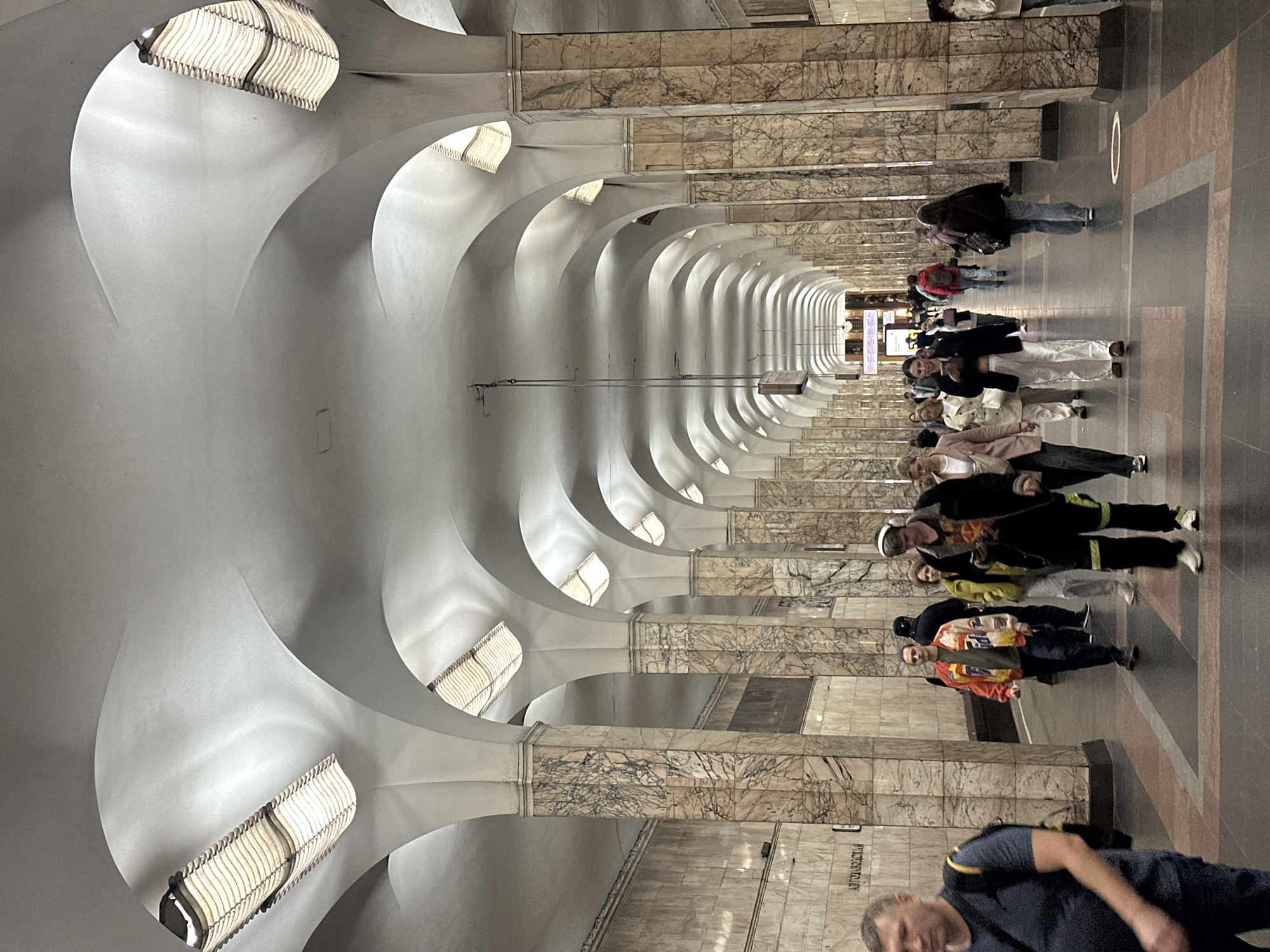 The image depicts the interior of a metro station in Moscow, Russia, likely Avtozavodskaya station, given the visible signage. The station features a grand, cavernous design with a series of interconnected arches forming a long, vaulted ceiling. Large, marbled columns line the walkway, supporting the arches and creating a repetitive architectural pattern that recedes into the distance. Rectangular light fixtures are embedded within the ceiling's curves, casting a soft, diffused light. The floor is paved with dark tiles, interspersed with reddish-brown rectangular sections, creating a geometric pattern.
Several people are walking in both directions along the platform. In the foreground on the left, a man wearing a dark blue t-shirt and a backpack walks towards the viewer. Further down the corridor, a group of individuals are seen walking away from the camera. These include a man in a colorful jacket, a woman in a white jacket, and others. Towards the right side of the image, more pedestrians are walking in both directions. One individual in the mid-ground on the right is wearing headphones and looking at their phone. The overall atmosphere suggests a typical day in a busy public transportation hub.