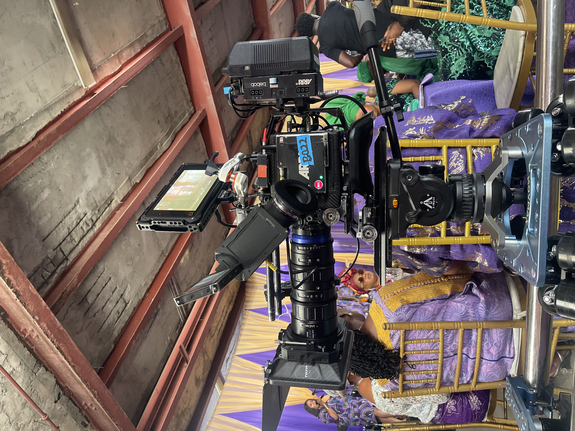 The image shows a professional camera setup in an indoor location, likely a church or event hall. The camera is mounted on a tripod and equipped with accessories like a monitor, lens controls, and an ARRI power distribution box. The camera points towards seated people who are dressed in what appear to be traditional African attire, primarily purple and gold colors. The event might be some kind of ceremony. The background includes golden chairs and a decorative purple and gold fabric draped on the seats. The ceiling of the room is visible and appears to be made of concrete and metal beams.