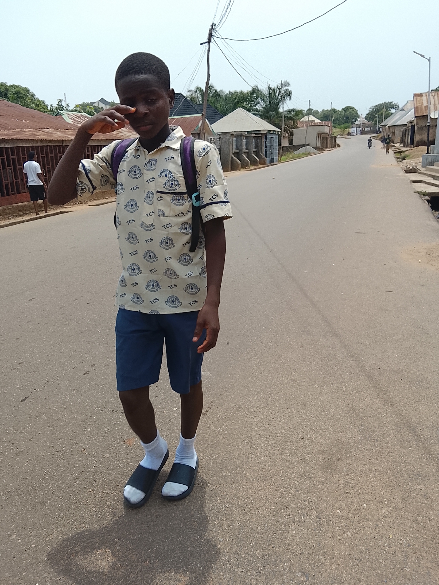 The image captures a young person walking down a seemingly quiet street in Akwanga, Nigeria, on a bright, sunny day. The primary subject, positioned in the foreground and center, is a young male with dark skin and short hair, appearing to be in his pre-teen or early teen years. He is wearing a patterned light-colored short-sleeved shirt, dark blue shorts, white socks, and black slide sandals. A dark purple backpack is visible on his shoulders. He is caught mid-action, rubbing his right eye with his hand, which could suggest tiredness, irritation, or an attempt to clear his vision. His expression is somewhat obscured but appears neutral or slightly weary.
Behind the main subject, another person in a white t-shirt and dark shorts is seen walking away on the left side of the street. The street itself appears to be paved with asphalt or a similar material, with unpaved shoulders and drainage channels on the sides.
The surrounding environment consists of residential buildings with corrugated metal roofs, many showing signs of weathering and rust, typical of some areas in Nigeria. Concrete block walls and fences are also visible. Utility poles with numerous overhead wires line the street, hinting at the infrastructure. Further down the road, the street curves gently, and more buildings and distant trees are visible under a clear, bright sky. A motorbike and a few individuals can be seen in the distance, suggesting typical daily activity.
The visible text on the main subject's shirt repeatedly displays "TCS" along with a circular logo containing what appears to be a crest or emblem. The overall scene conveys an ordinary day in a residential area, characterized by its sunny conditions and rustic architectural style.