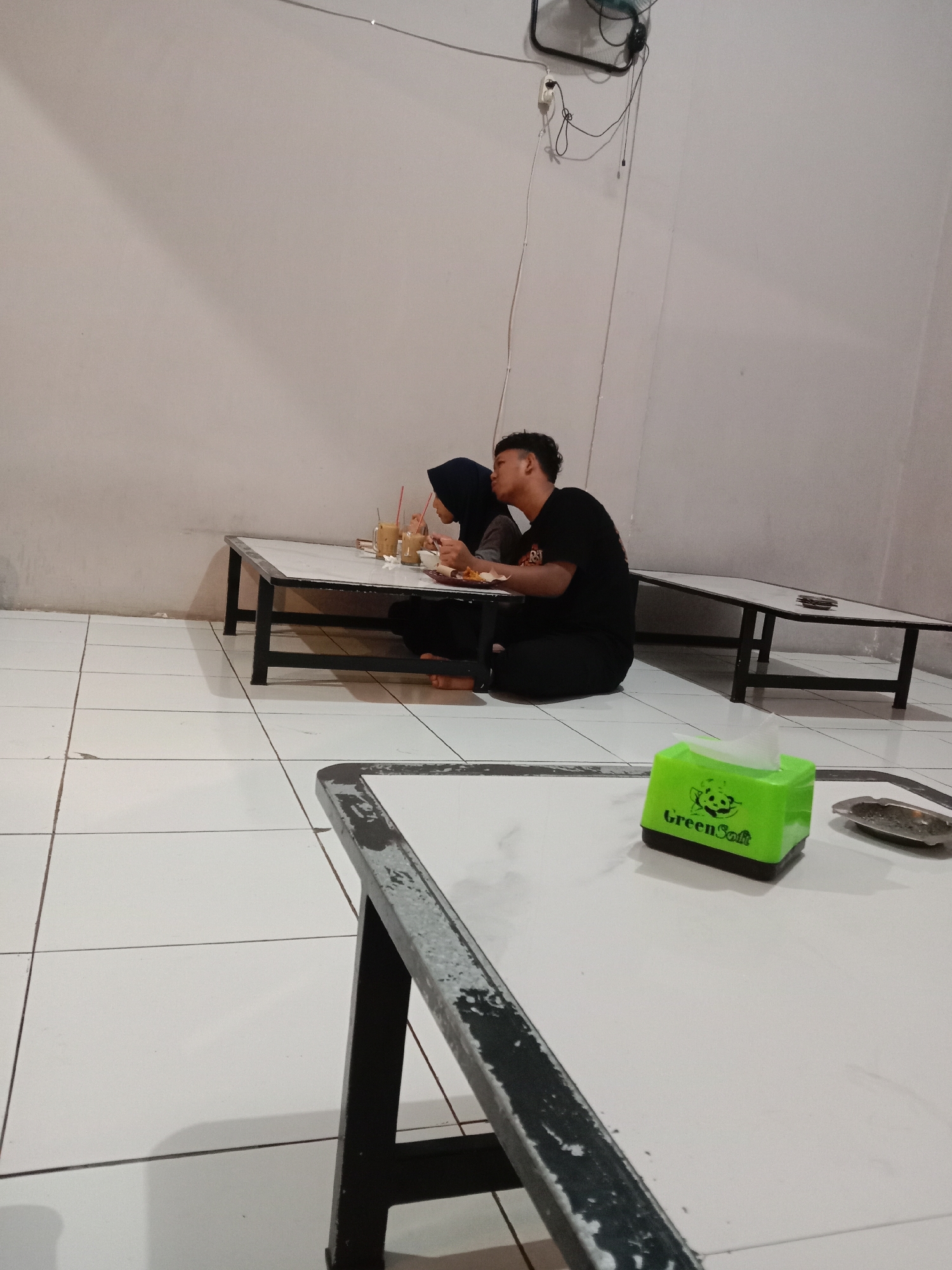 A couple is seated at a low table in what appears to be a restaurant or food stall setting in Cangkring, Indonesia. The woman is wearing a hijab, while the man is in a black t-shirt and pants. They are sharing a meal and drinks. A green tissue box sits on a table in the foreground. The floor is tiled. A wall fan is visible.
