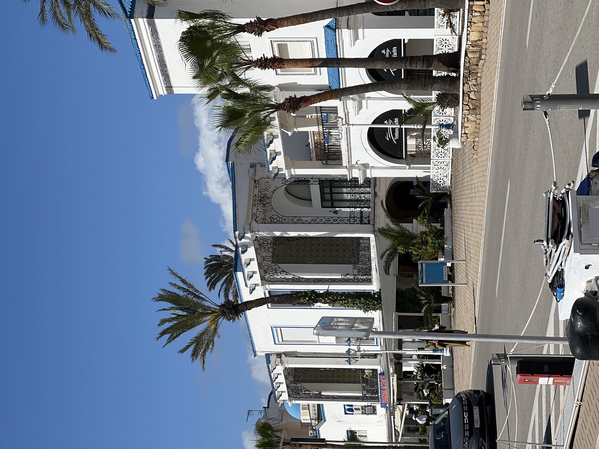 The image shows a street scene in M'Diq, Morocco, characterized by white buildings with blue accents, a style typical of the region. Palm trees line the street, adding to the coastal atmosphere. The architecture features arched doorways and decorative ironwork balconies. A road runs alongside the buildings, with vehicles parked along the side. There are signs visible, indicating commercial establishments. A mix of residential and commercial elements is suggested by the presence of both balconies and business signage. The sky is a clear blue with a few clouds.