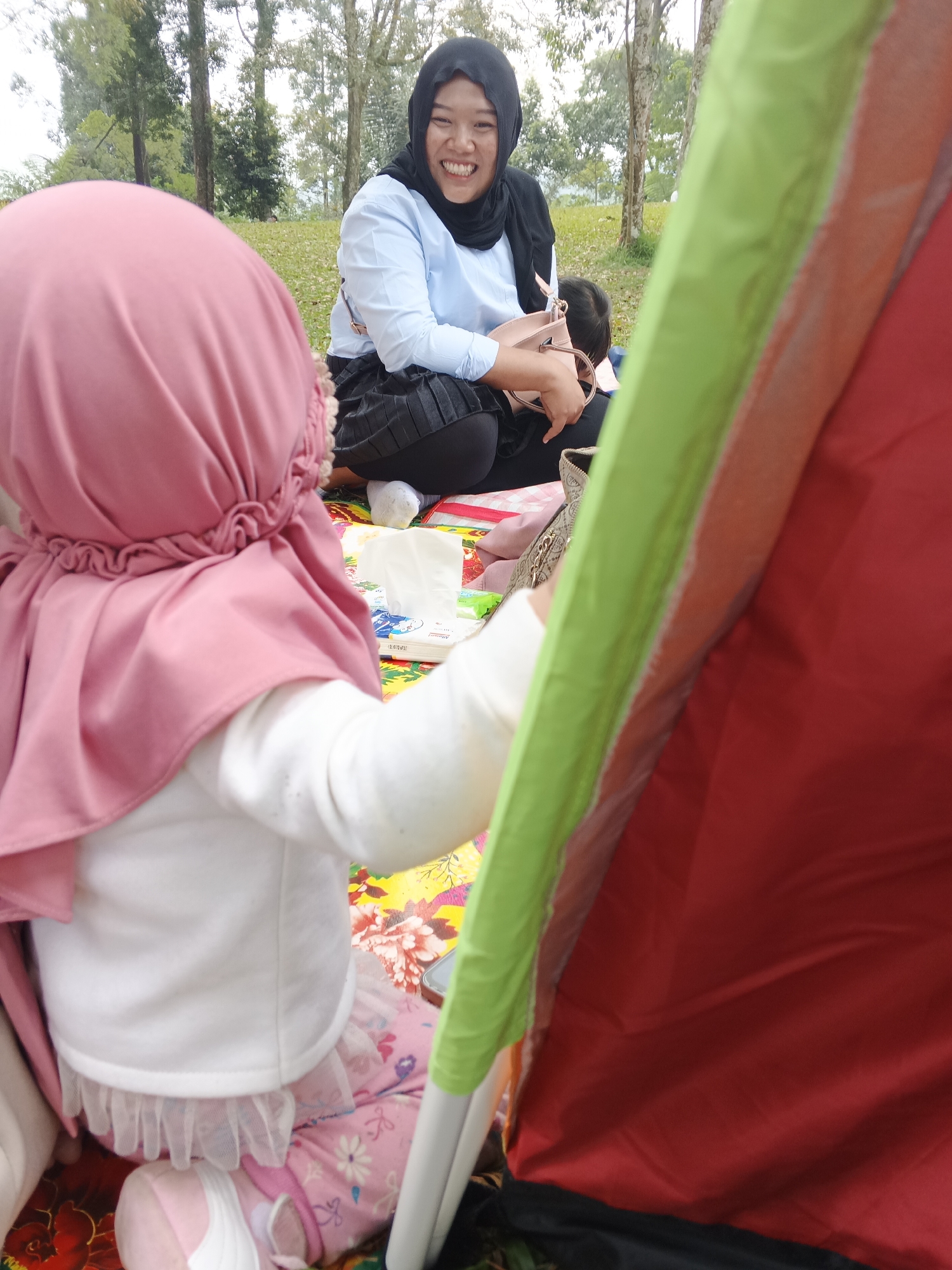 In Perbawati, Indonesia, a woman wearing a black hijab and light blue shirt smiles broadly while holding a pink purse. She is seated on a colorful mat outdoors, with a young child in a pink hijab and white shirt facing away from the camera. The child is wearing pink pants with a floral pattern and a white tutu-like underskirt. A tent with red and green panels is partially visible in the foreground, obscuring some of the scene. In the background, several trees with sparse foliage are visible, suggesting a park or forested area. The ground is covered with grass and scattered leaves. A pack of tissues is placed on the mat near the child.