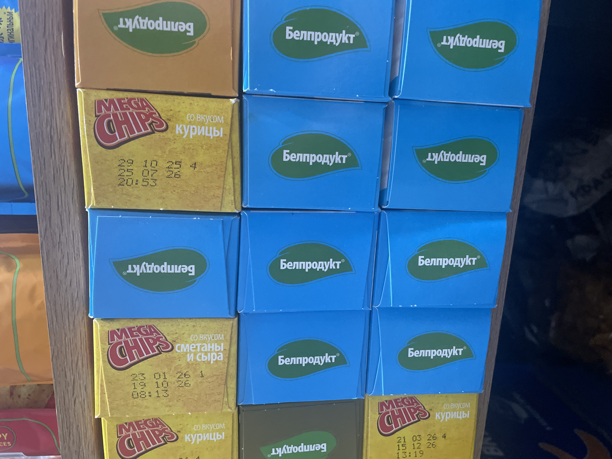 The image shows a retail shelf displaying several rows of packaged snack products. The products are predominantly small cardboard boxes. In the top visible row, there are yellow and orange boxes with text and logos. The yellow boxes appear to have "MEGA CHIPS" printed on them, along with flavor descriptions in Russian such as "сметаны и сыра" (sour cream and cheese) and "курицы" (chicken). Some boxes have date codes printed on them, for example, "21.03.26".
Below the top row are rows of bright blue boxes, each featuring a green leaf logo and the Russian text "Белпродукт" (Belprodukt). These blue boxes are arranged in multiple columns. The lighting suggests an indoor retail environment. The shelf itself appears to be made of wood. The overall scene is that of a product display in a store, likely a convenience store or a small grocery shop. The city of Zhukovo, Russia, is mentioned as a location but there are no visual cues in the image that confirm or deny this specific location.