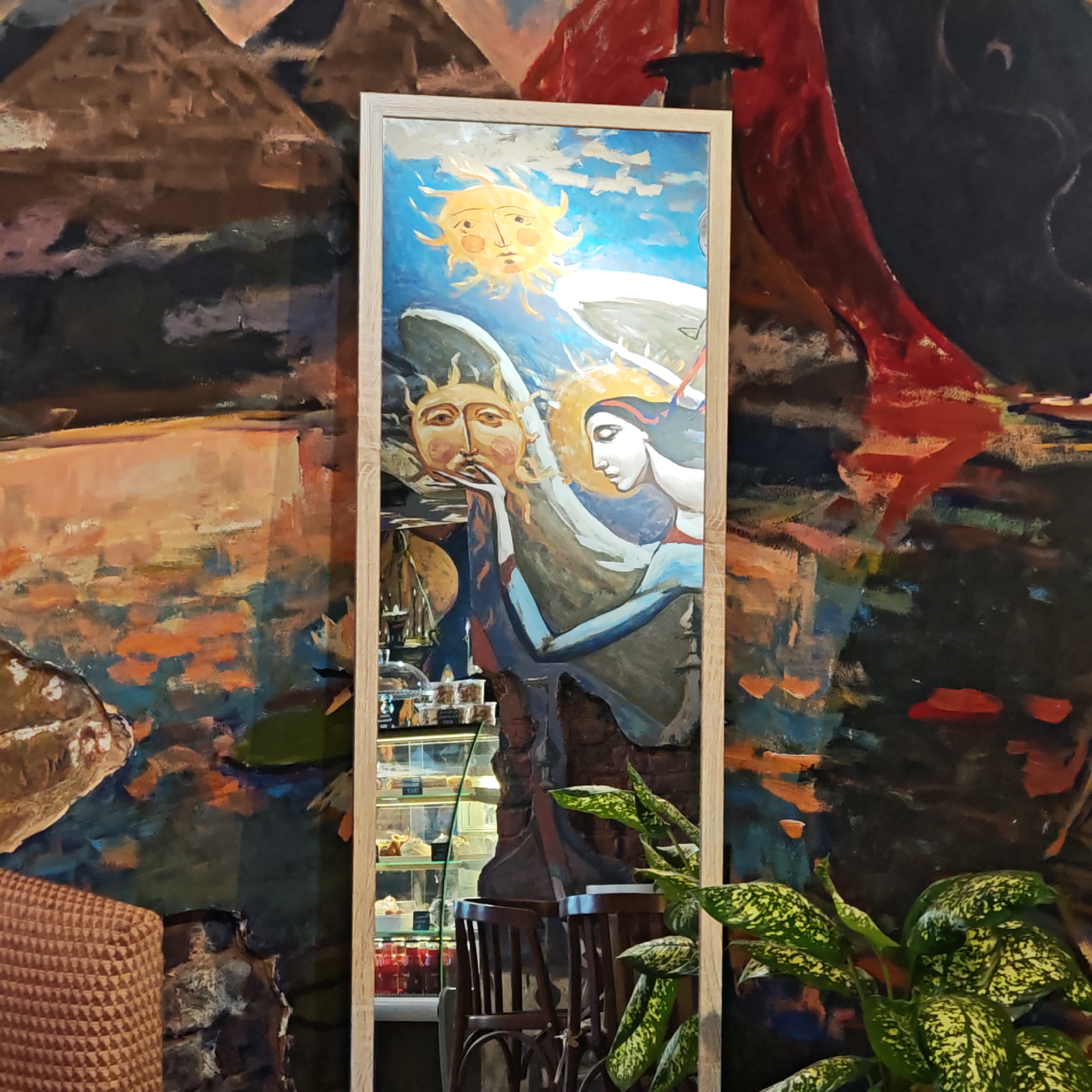 The image is a view of an interior, likely in a public place such as a cafe, in Rostov, Russia. The focal point is a framed artwork of a somewhat folk-art style depicting two sun-like figures, one with a human face, and a female figure with a halo. The artwork is vertically oriented and appears to be hanging on a wall. Reflections in the frame reveal part of a display case filled with pastries or food items, and wooden chairs are nearby. A potted plant with speckled green and yellow leaves is located on the right side of the image. The background wall features an abstract painting with browns, blacks, reds, and oranges, giving a somewhat textured or landscape-like impression. Part of a patterned chair or cushion is visible in the lower left corner. The overall lighting is soft and diffused, creating a somewhat warm and inviting atmosphere.