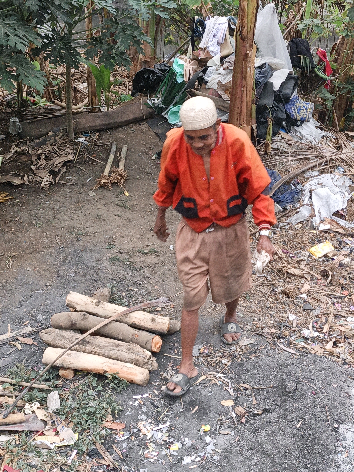 An elderly man wearing a white skullcap and an orange jacket over tan shorts walks through an area strewn with debris, suggesting an urban fringe or informal settlement. He is looking down at something in his left hand, which appears to be a piece of white cloth. He wears dark sandals with a star emblem. In the foreground, several logs are stacked on the ground, and a long, thin branch lies across them. The background is cluttered with piles of trash, including plastic bags, clothing, and various types of refuse, amidst vegetation that includes banana plants. The ground is a mixture of dirt and ash. The overall impression is one of poverty and the presence of accumulated waste. The location is identified as the city of Mangli, Indonesia.