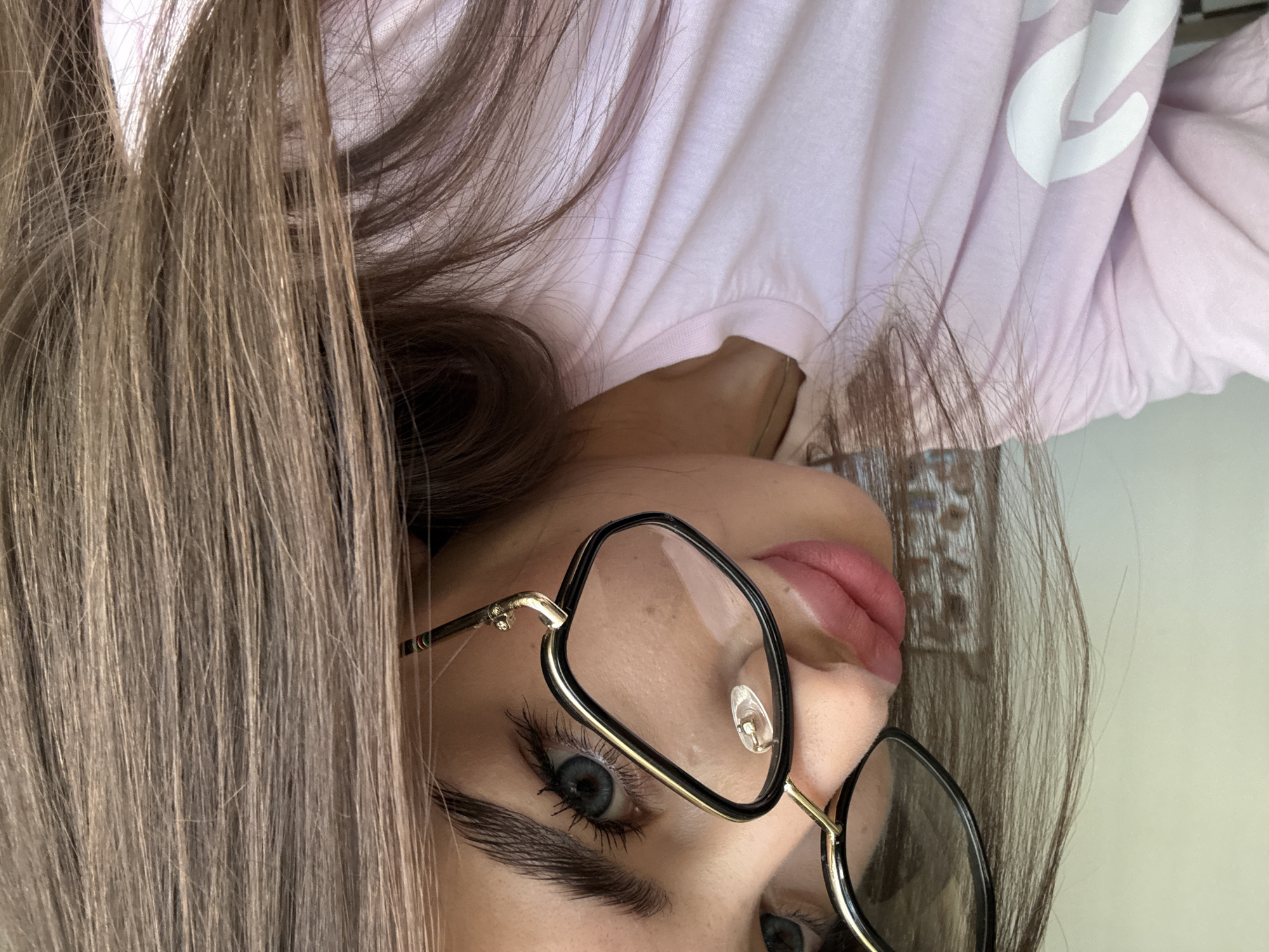 The image is a close-up, selfie-style shot of a young woman with blue eyes and light brown hair. She is wearing large, black-rimmed glasses with gold accents and a light pink t-shirt with a white graphic on it. The background is a plain, light-colored wall. There is no discernible information about the location in Chișinău, Moldova. The woman is looking slightly to her left, with her lips slightly parted. The lighting suggests an indoor setting.