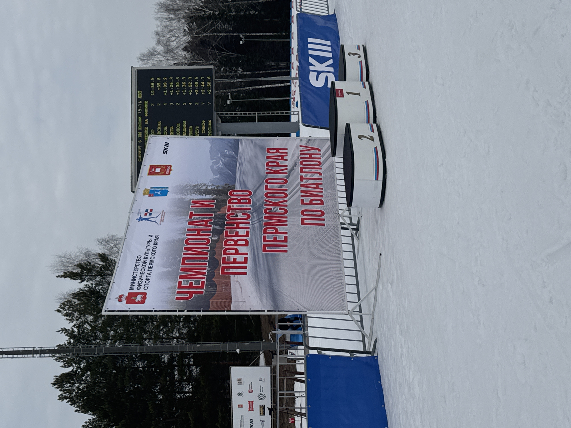 This image depicts an outdoor winter scene at a biathlon competition venue in Прикамский, Perm Krai, Russia. The foreground is covered in white snow, leading to a three-tiered podium for awards, numbered 1, 2, and 3, featuring white bases with red, blue, and white stripes, and black tops. Behind the podium, a large white banner is prominently displayed, stating "ЧЕМПИОНАТ И ПЕРВЕНСТВО ПЕРМСКОГО КРАЯ ПО БИАТЛОНУ" (Championship and Championship of Perm Krai in Biathlon) in red Cyrillic text. This banner also includes logos for the Ministry of Physical Culture and Sports of Perm Krai, the Perm Krai coat of arms, and "SKIII." To the right of the main banner, an electronic scoreboard displays competition results, reading "СПРИНТ 6 КМ ЮНОШИ 15-16 ЛЕТ" (Sprint 6 km Boys 15-16 Years) and "ПОЛОЖЕНИЕ НА ФИНИШЕ" (Position at Finish), along with a list of names and times. In the background, a dense line of snow-dusted coniferous and deciduous trees stands under an overcast sky. Metal barriers and a blue "SKIII" banner are visible behind the podium and along the perimeter. A tall metal lattice tower is present on the far left. No individuals are visible in the scene.