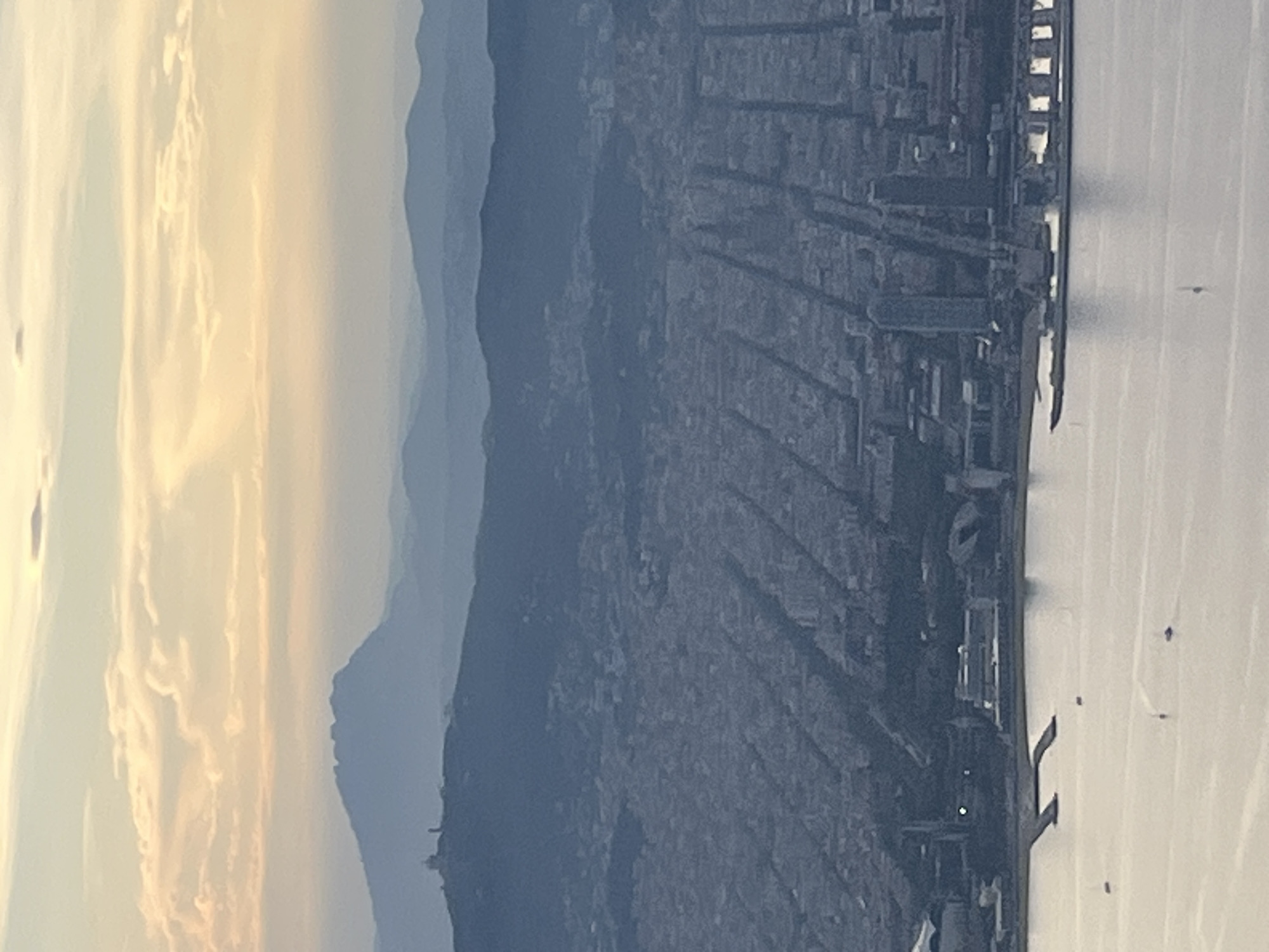 The image shows an elevated view of Barcelona, Spain, at what appears to be dusk or dawn. To the left are the mountains surrounding the city. The city grid with its dense buildings is visible in the center. To the right is the Mediterranean Sea. The scene shows no people visible in the frame.