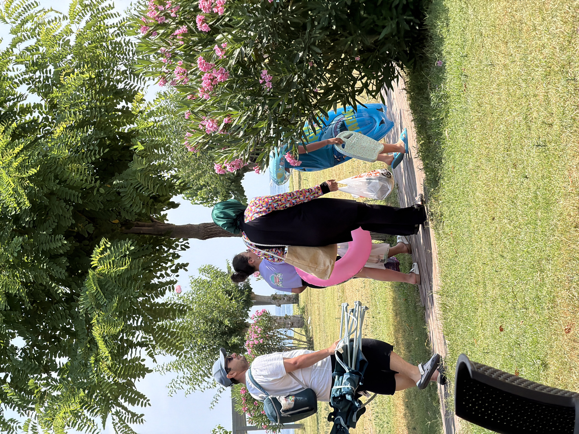 A group of people are walking on a paved path next to a grassy area with trees and flowering bushes. The location appears to be a park or outdoor recreational area in Kemer, Turkey, with the Mediterranean Sea visible in the background.
In the foreground, a man wearing a white t-shirt, black shorts, and a blue baseball cap with sunglasses is carrying a cooler bag on his back and a folded beach chair or umbrella in his right arm. He is wearing grey crocs. To his left, a person is partially visible in a black chair.
Behind the man, three other individuals are walking. One is a young woman in a purple t-shirt and dark shorts, holding a pink inflatable ring. Next to her, a woman is wearing a black full-body swimsuit with a colorful patterned neckline and sleeve accents. She has a green headscarf and is carrying a beige tote bag and a white plastic bag. To her right, a child is wearing a blue t-shirt with shark graphics and blue shorts. The child is carrying a light blue inflatable ring and a pale blue mat or tray.
The environment features lush green trees with feathery leaves, vibrant pink oleander bushes in bloom, and a clear blue sky. The paved path is made of light-colored stones. The overall scene suggests a warm, sunny day suitable for outdoor activities.