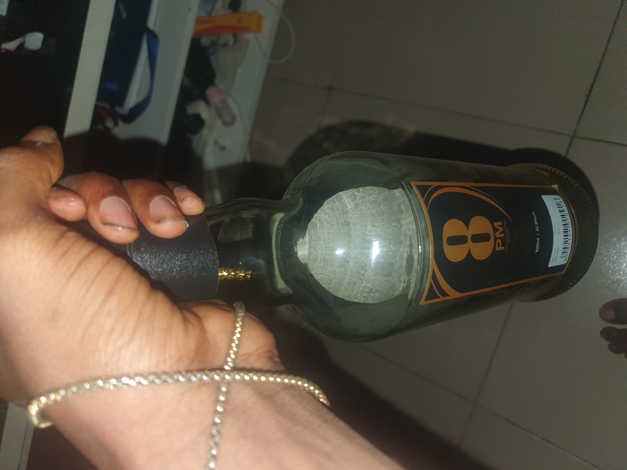 A person is holding a nearly empty bottle of 8 PM Grain Blended Whisky. The bottle is held horizontally. The person has dark skin and is wearing a silver bracelet. The bottle has a black label with the number "8" in gold. The bottle is 1000ml and 42.8% alcohol by volume. The background is blurry but appears to be a tiled floor and some objects on a shelf. The exact location in Lokoja, Nigeria is not discernible from the image.