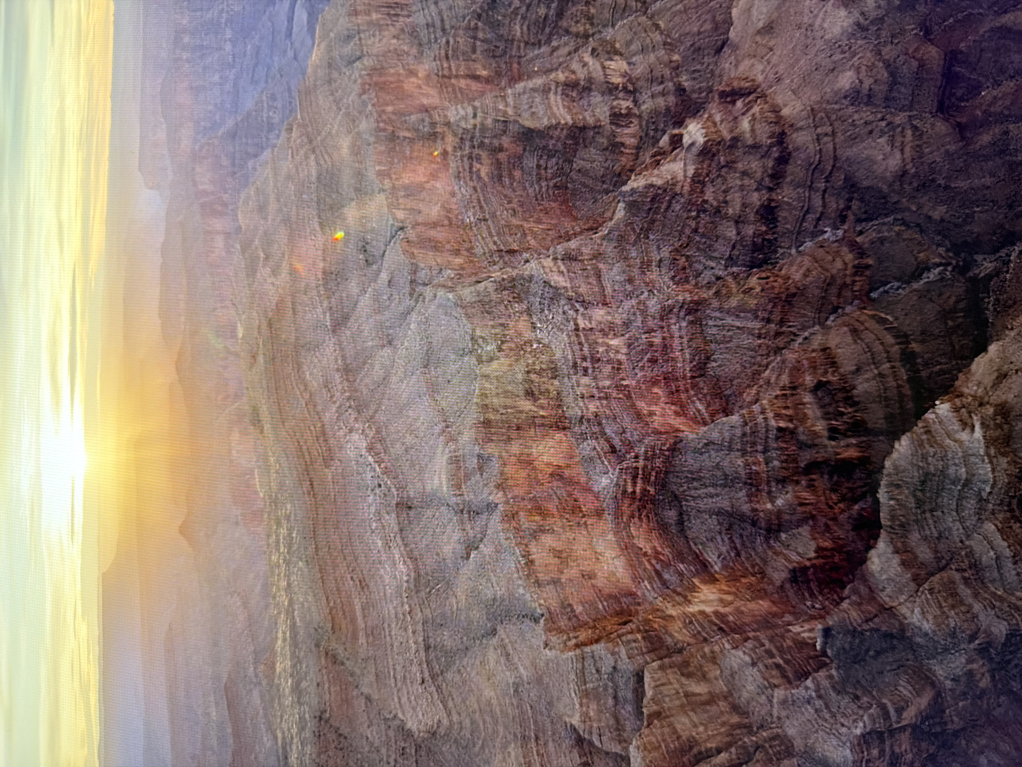 The image shows an aerial view of a vast canyon landscape during a sunset. The sun's rays are visible, casting a warm glow on the layered rock formations. The canyon walls display distinct strata of varying colors, ranging from earthy browns and reds to purples and grays. In the distance, hazy mountain ranges are visible under a pale sky. The perspective suggests a high vantage point, possibly from an aircraft. There are no people, objects, or discernible structures that would indicate a location in Kyiv, Ukraine. The scene is entirely natural and geological.