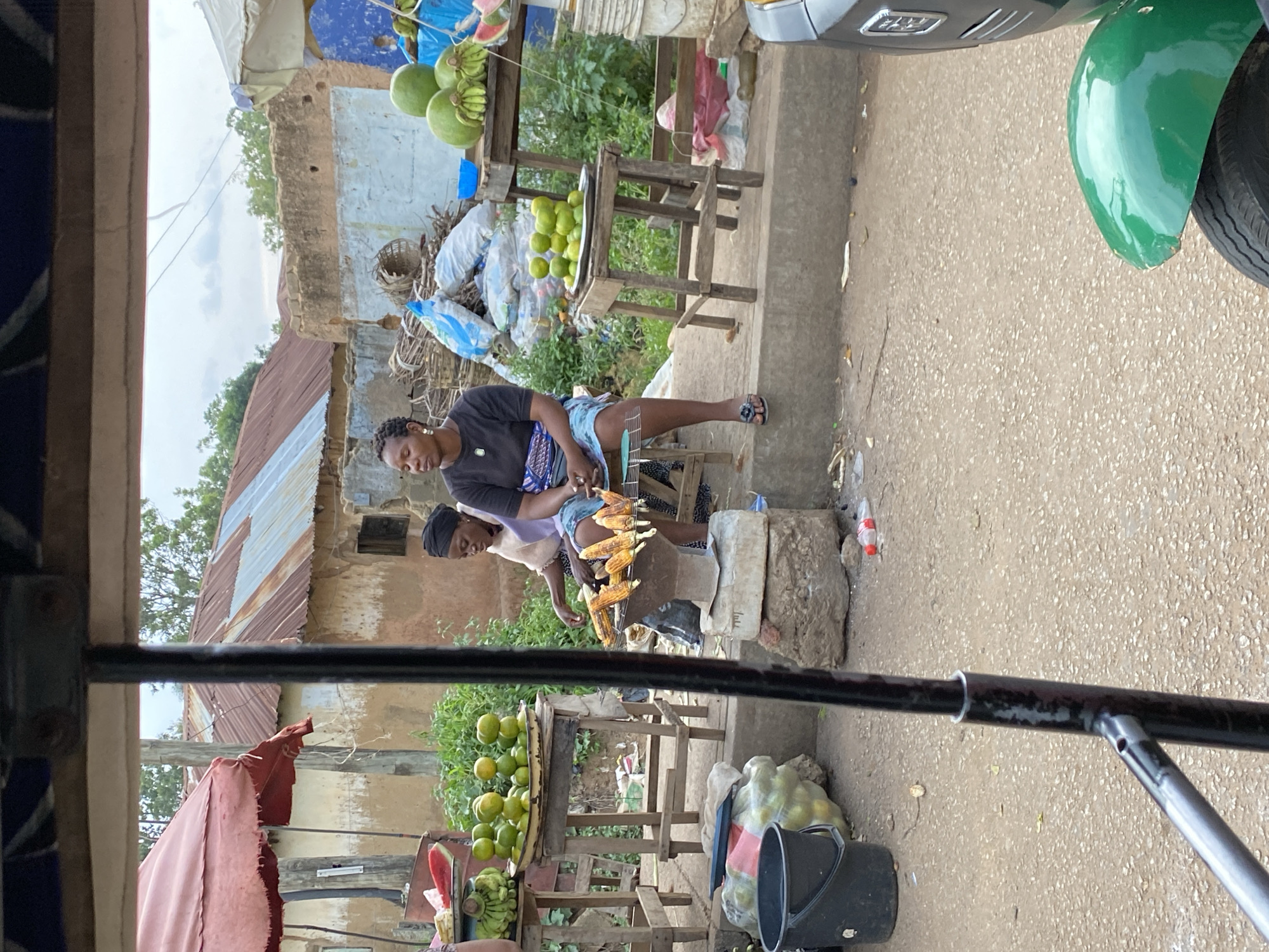 A street scene in Bukuru, Nigeria, shows a market stall with produce and two women. A person is seated on a stool and selling corn on the cob, which is displayed on a metal stand. Another woman is seated behind the corn seller. In the foreground, a black bucket and a pile of green fruit are visible. The background features several buildings with corrugated metal roofs, as well as some greenery. A portion of a green vehicle is visible in the lower right corner of the image.