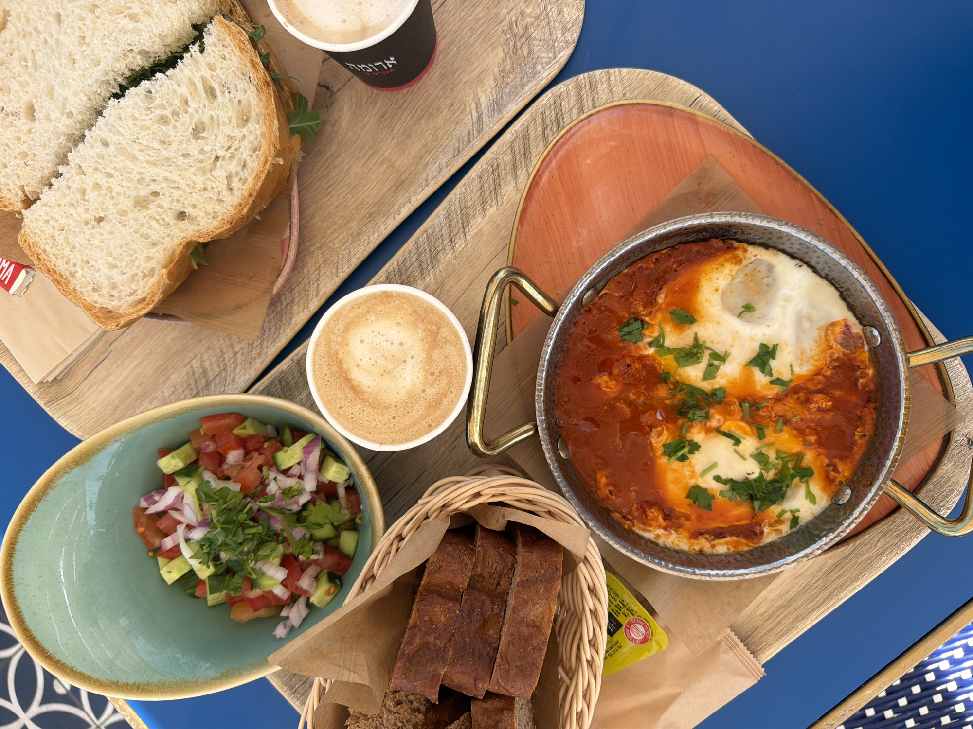 The image shows an overhead shot of a breakfast spread on a blue table, likely in a Tel-Aviv, Israel cafe setting. The spread includes a sliced sandwich on light bread, accompanied by a cup of coffee in a branded cup. Another cup of coffee is also visible near a bowl of chopped cucumber and tomato salad, which features some red onion and herbs. There's a basket of dark bread slices. Also, a pan of shakshuka, or eggs in tomato sauce, is present. A small packet of butter is seen near the shakshuka and bread. Everything is arranged on wooden serving trays.
