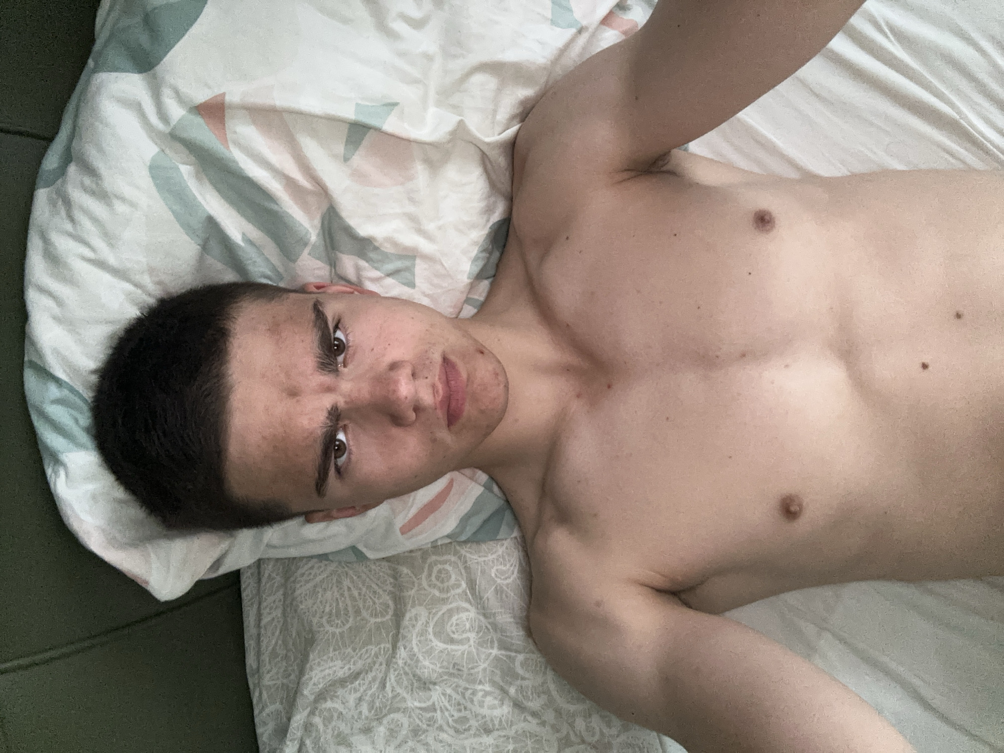 A young man with light skin, short dark hair, dark eyebrows, and brown eyes lies shirtless on a bed or sofa, looking directly into the camera. He is positioned on his back with his head resting on a white pillow adorned with a pastel geometric pattern in shades of green, pink, and grey. His body is lean with visible chest muscles, and small moles or freckles are scattered across his torso. His facial expression is somewhat intense, with slightly furrowed eyebrows and a closed mouth that gives a serious or slightly pouty look. Visible blemishes are present on his forehead and chin.
One arm is raised and partially visible towards the top of the frame, suggesting he might be holding the camera for a selfie. The bedding beneath him is white with a subtle grey pattern, and a dark green surface, possibly a headboard or part of a furniture piece, is visible to the left. The lighting is soft and even, without harsh shadows. The setting appears to be an indoor personal space, likely a bedroom. There is no visible text in the image.