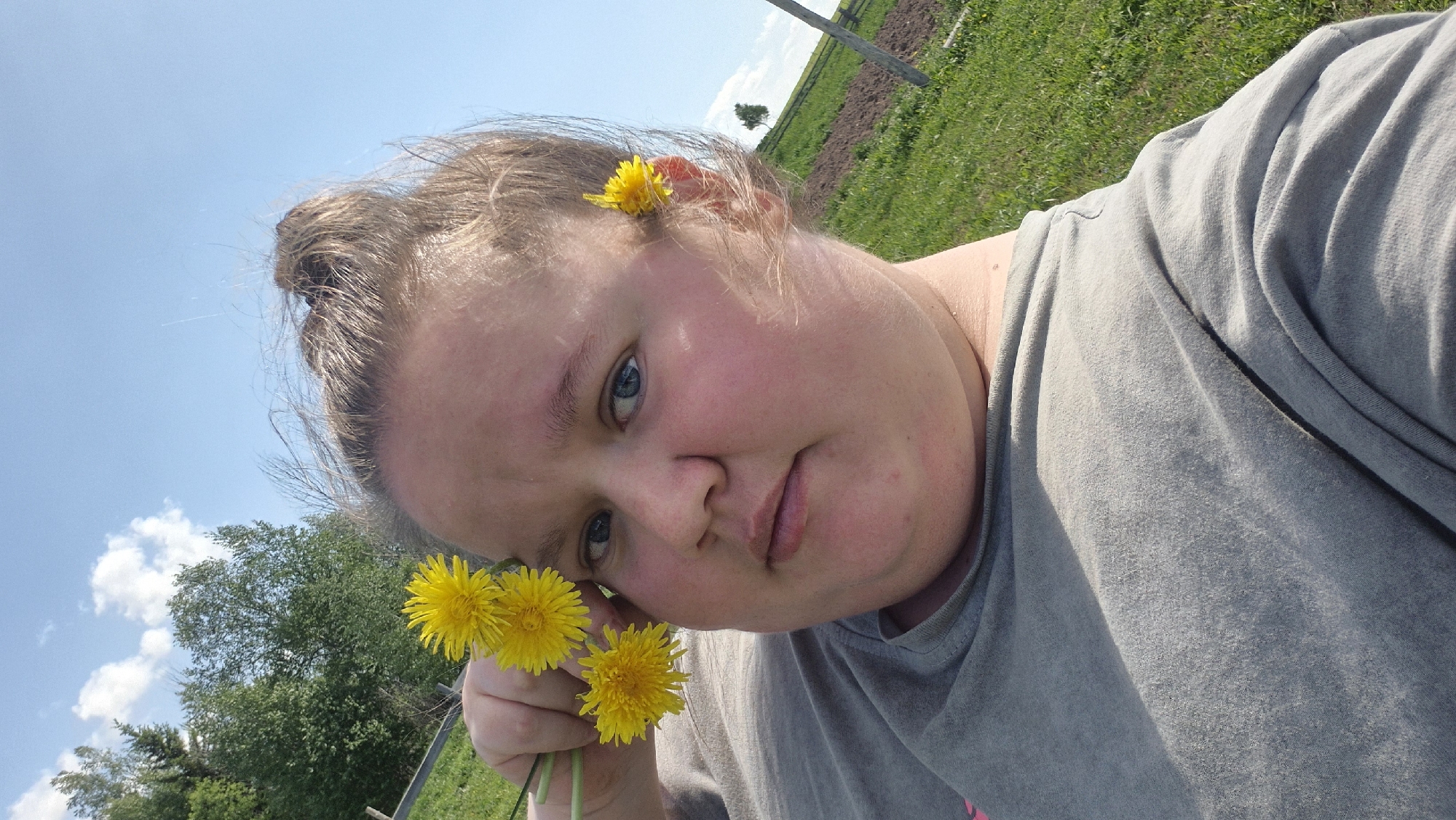 A woman is holding dandelions. One dandelion is in her hair. The location is Дойная, Russia. The background has grass and the sky. The woman is wearing a gray shirt.