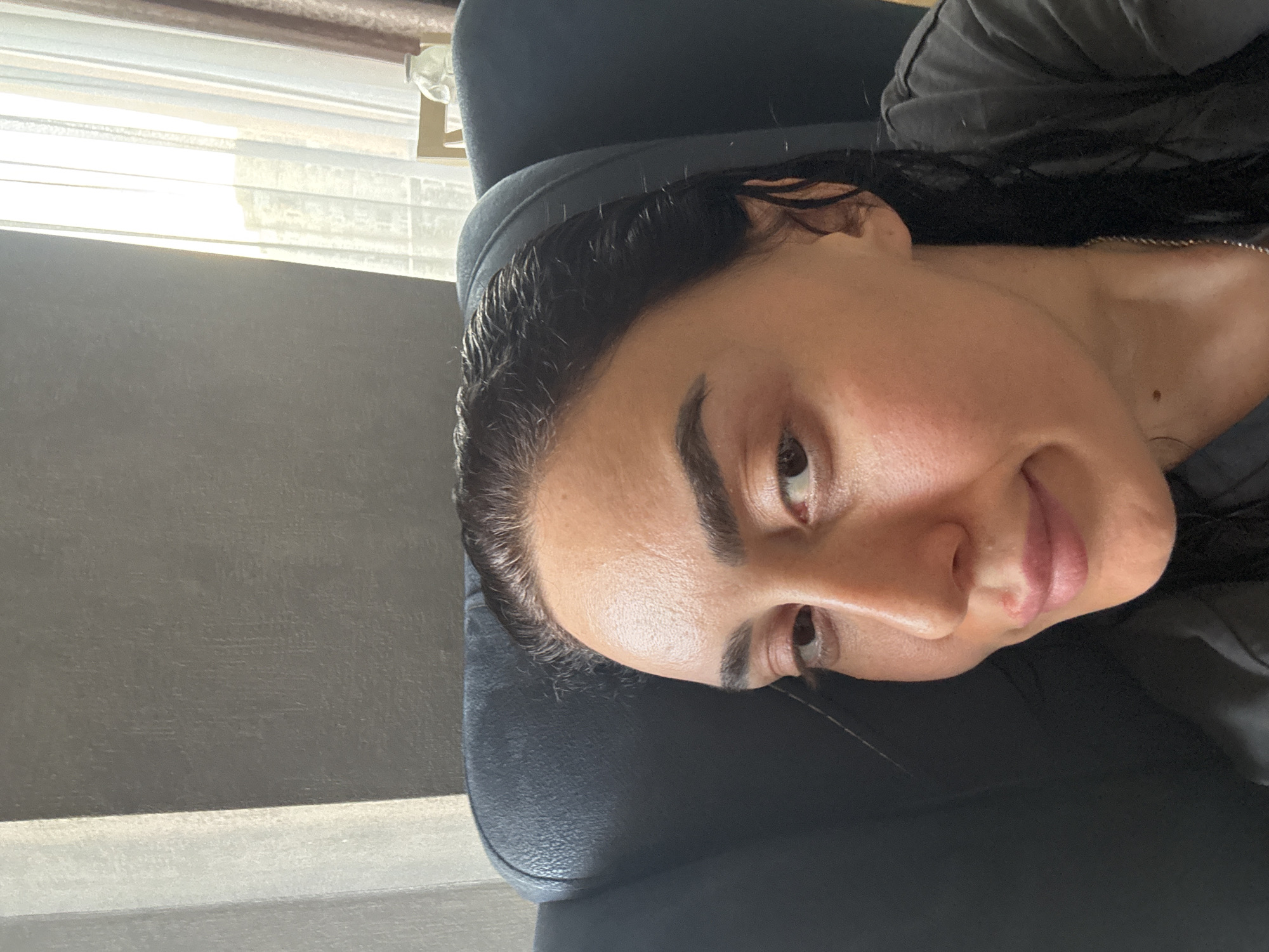 The original image, captured in Tbilisi, Georgia, is rotated 90 degrees counter-clockwise. When viewed correctly, it presents a close-up, waist-up portrait of an adult woman. She is positioned slightly to her left, looking towards the camera with a calm expression and a subtle, closed-mouth smile. Her dark hair is styled back from her face, complementing her dark eyebrows and eyes. Her skin shows a natural sheen, likely from the bright, natural light illuminating her from the left. She wears a dark grey or black casual top, with a delicate silver-colored necklace around her neck. A small blemish is visible on her upper lip. The woman appears to be leaning against a dark, possibly leather, chair or headrest. In the background, bright natural light streams through a window fitted with white horizontal blinds, visible on the left side of the frame. The remainder of the background consists of a plain dark gray wall. No text is visible.