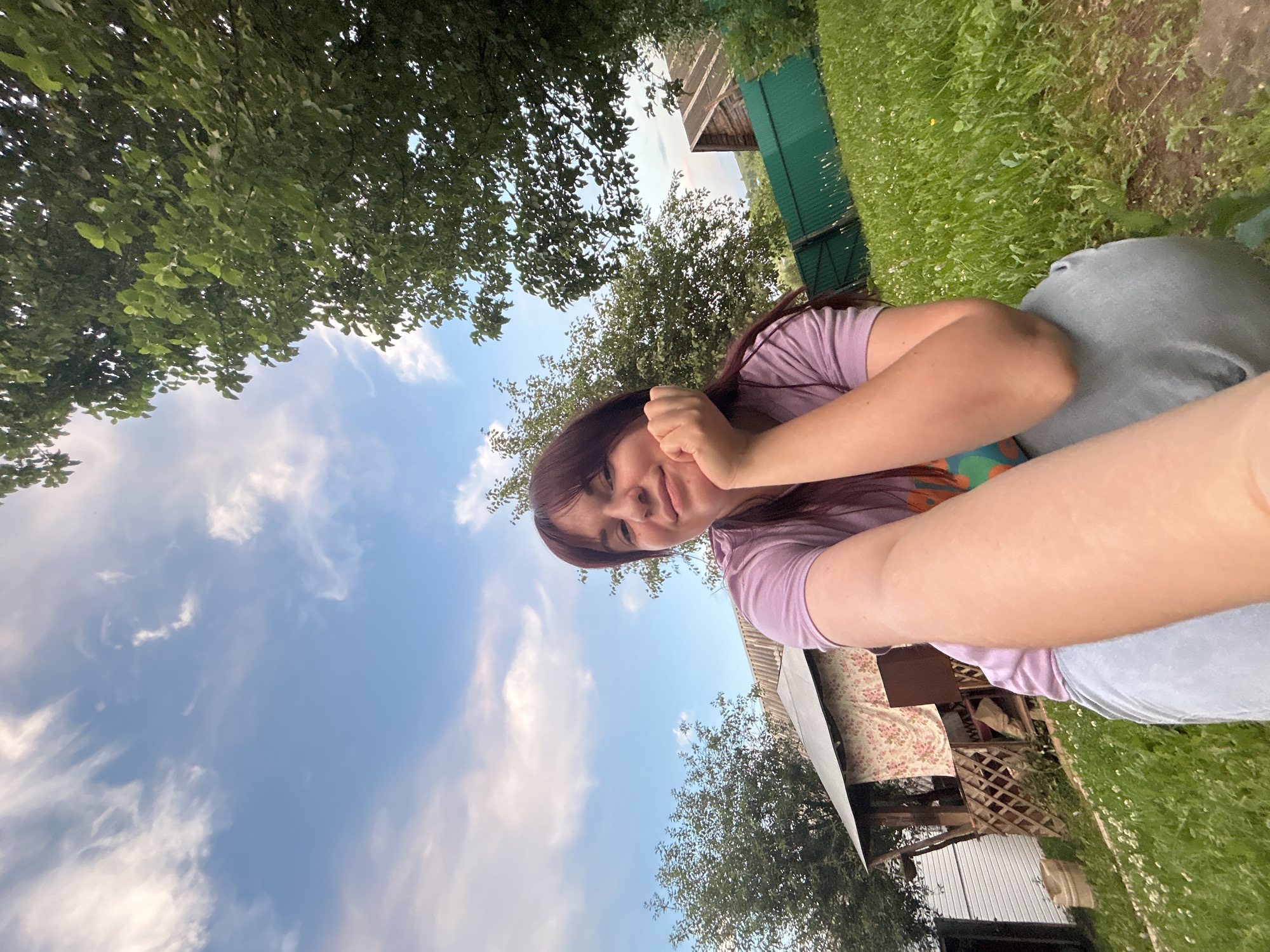 A young woman with long, dark hair is captured in a selfie-style photograph outdoors, with a backdrop of a rural residential area. She is wearing a light purple t-shirt and light-colored pants. Her right hand is resting on her chin, and she is looking towards the camera with a slight smile.
The environment appears to be a yard with a patch of green grass in the foreground, scattered with small white flowers. Behind the woman, there is a small house with a porch, a wooden fence, and a green fence. Trees with green foliage are visible on either side of the frame, and the sky above is a clear blue with scattered white clouds. The overall setting suggests a casual, outdoor scene in a village or suburban area, possibly the city of Фоки, Russia, given the context. The image is taken from a low angle, looking up towards the woman and the sky.