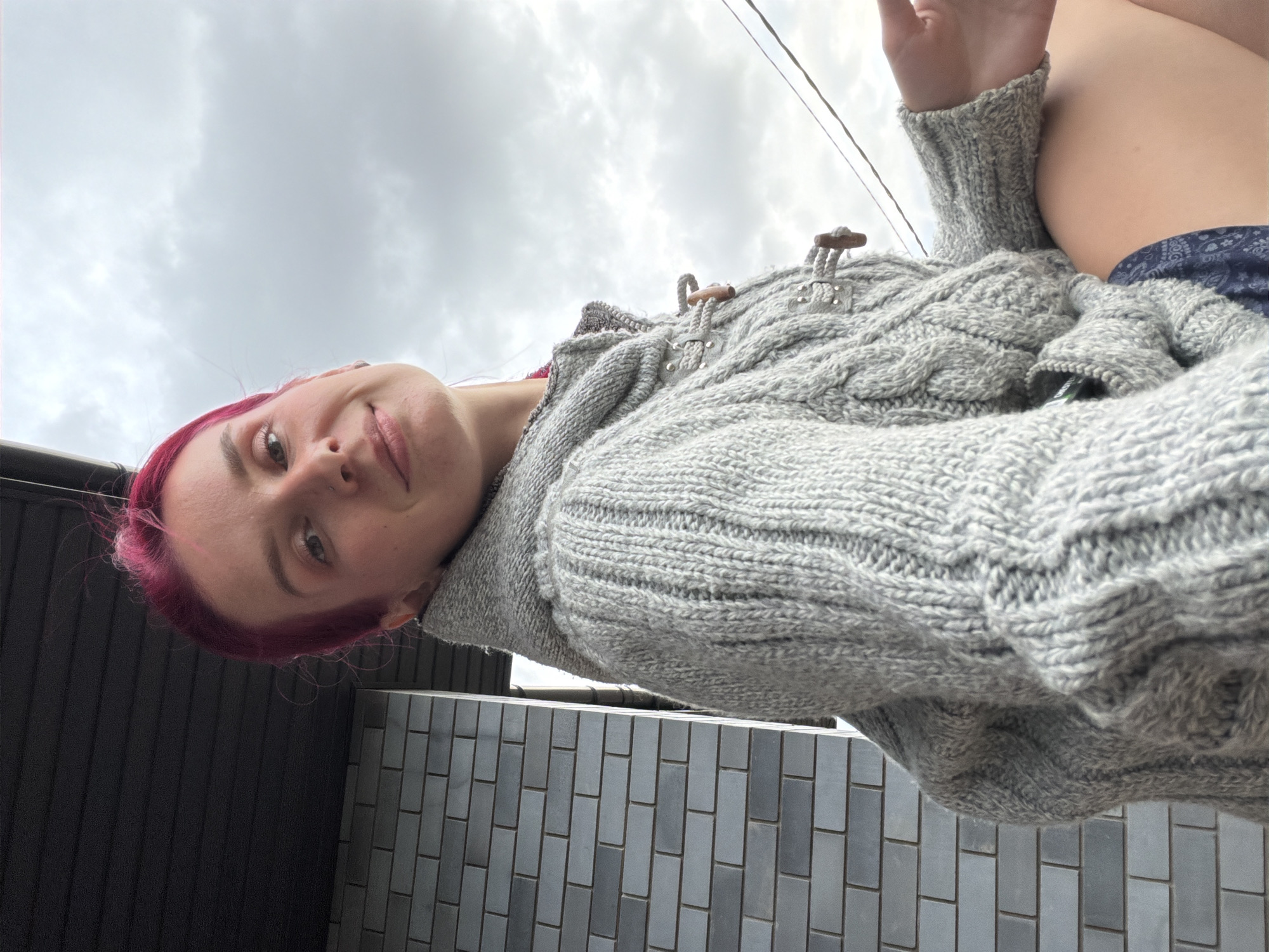 A woman with bright pink hair is shown from the chest up, leaning towards the camera. She is wearing a grey knit sweater with a hood and toggle closures. She has a slight smile and her eyes are looking at the camera. Behind her, a gray brick wall is visible, with a dark overhang above it. The sky, which is overcast and cloudy, fills the background. The image appears to be taken outdoors, possibly on a balcony or porch, with hints of an urban environment.