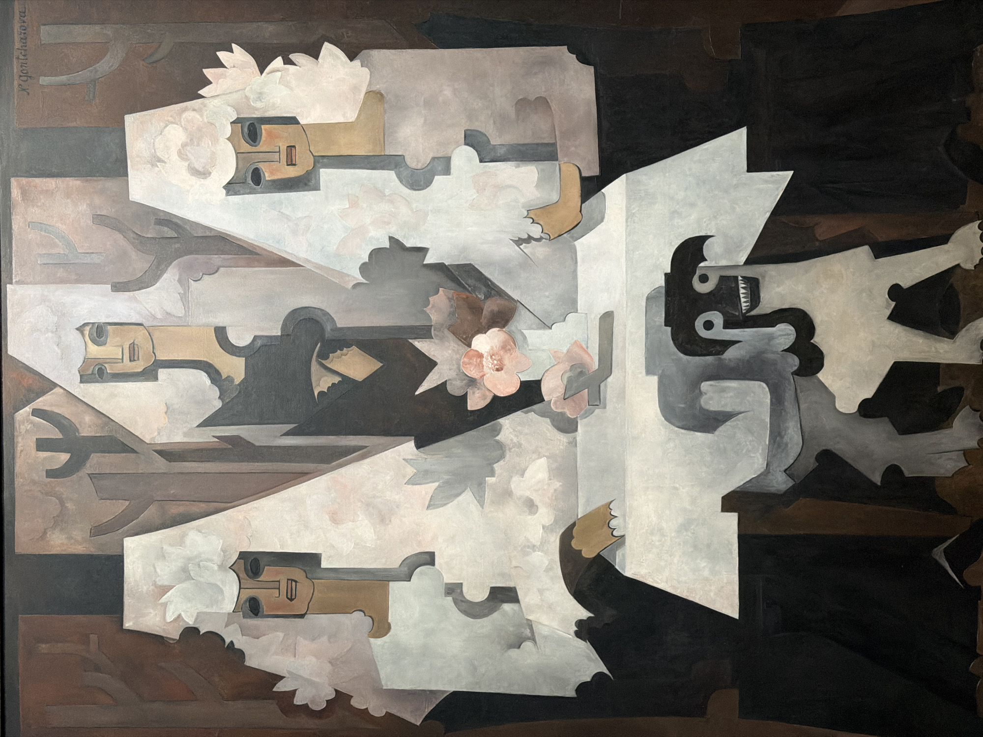 The image depicts an abstract painting with Cubist elements, likely created in Malakhovka, Russia. Three stylized, elongated human figures with simplified facial features and brown skin tones are positioned horizontally on the left side. Each figure has a white floral or cloud-like form emanating from the head. Grey and brown geometric shapes, possibly representing architecture or landscapes, surround the figures. In the upper left corner, a stylized signature "N. Goncharova" is visible. On the right side, a single figure in shades of grey and black is depicted, with sharp angular forms suggestive of a stylized animal or demon-like head with teeth, sitting or hunched over with implied motion. The background is composed of dark brown and black shapes, creating a sense of depth and shadow. A few muted pink flowers are scattered centrally in front of the figures on the left.