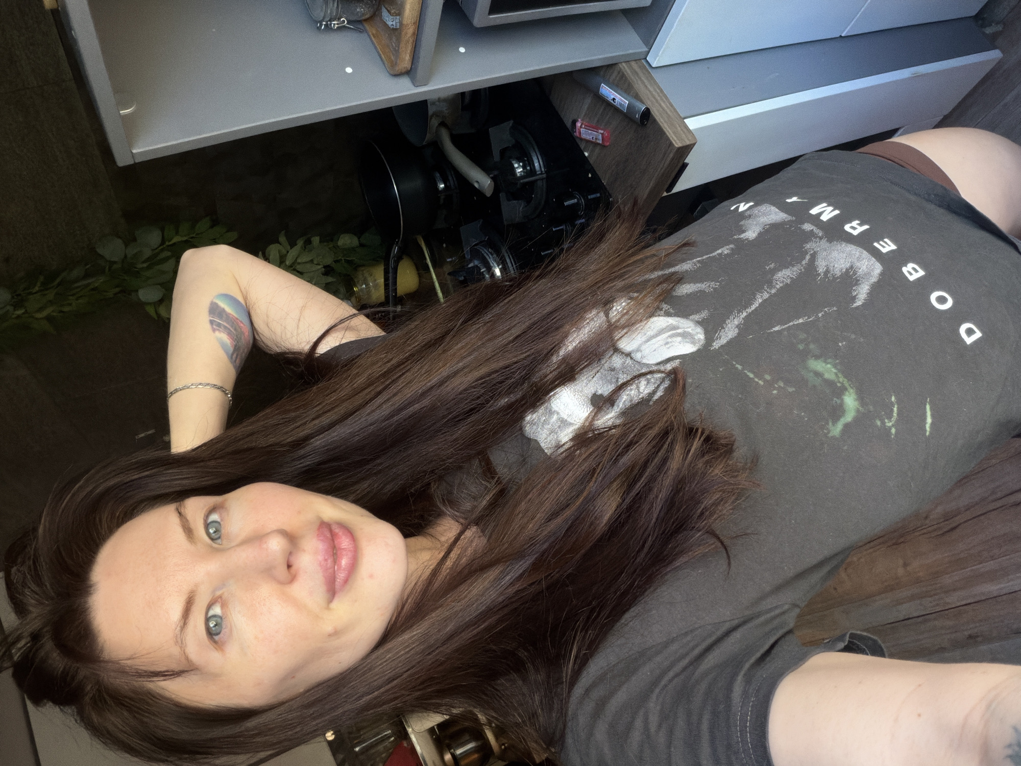 A selfie angled down showing a woman with light skin, blue eyes, and long dark brown hair, wearing a gray t-shirt with "DOBERMAN" printed on it, posing near an open drawer containing cookware, likely in a kitchen in a home or apartment. The exact location in Perm, Russia, is impossible to determine without additional information.