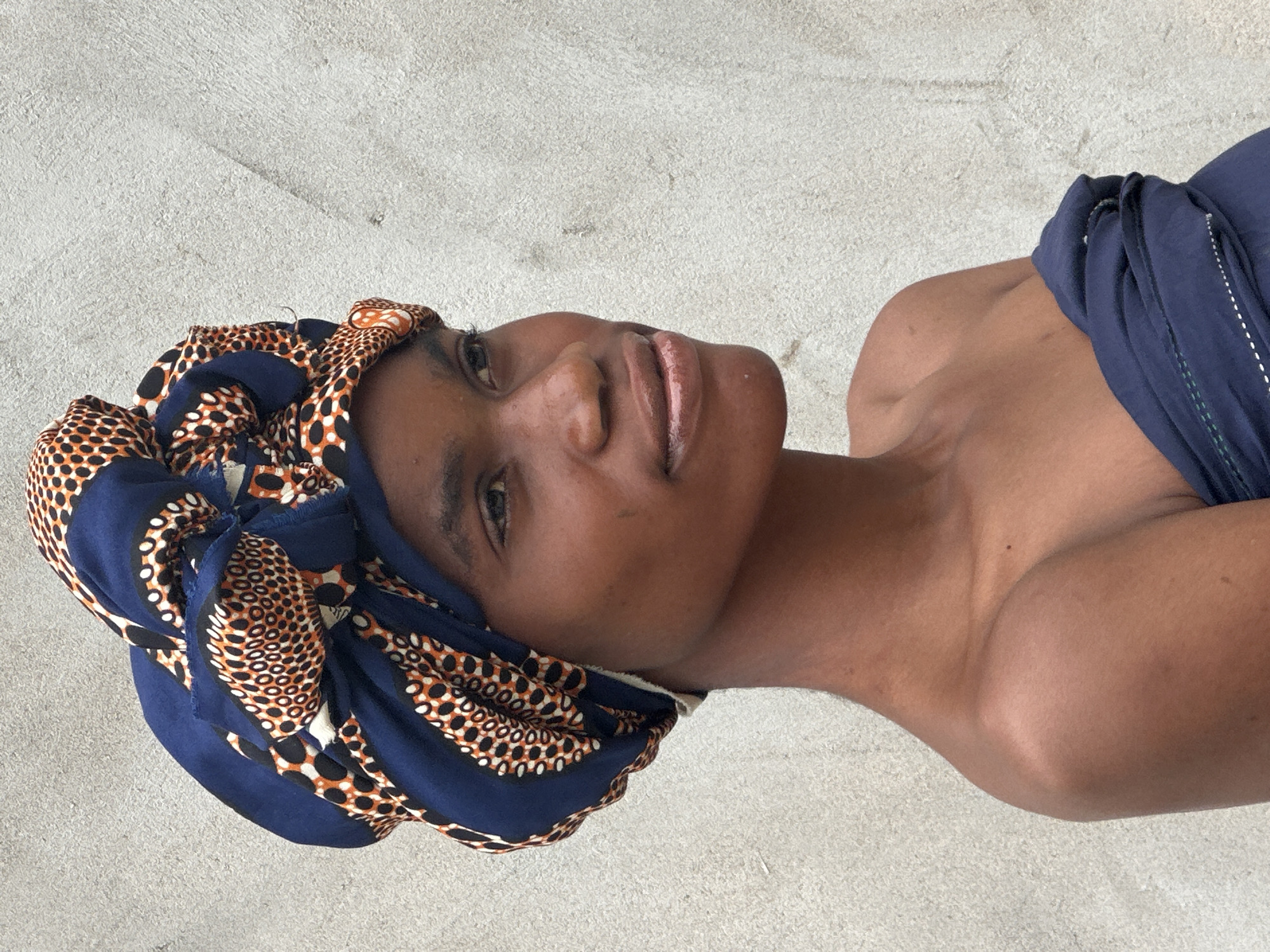 A medium shot captures a young Black woman from the chest up, positioned slightly off-center to the left. She is looking to her right, with her head tilted slightly upwards. Her face is turned in profile, revealing a smooth complexion and subtle makeup on her lips. She wears a vibrant, patterned headwrap that encircles her head, featuring bold geometric designs in navy blue, white, and orange. Her shoulders and upper chest are bare, with a dark blue fabric draped across her chest. The background is a textured, light gray wall. The image appears to have been taken outdoors, possibly in the city of Badagry, Nigeria, given the style of dress and the ambient lighting.